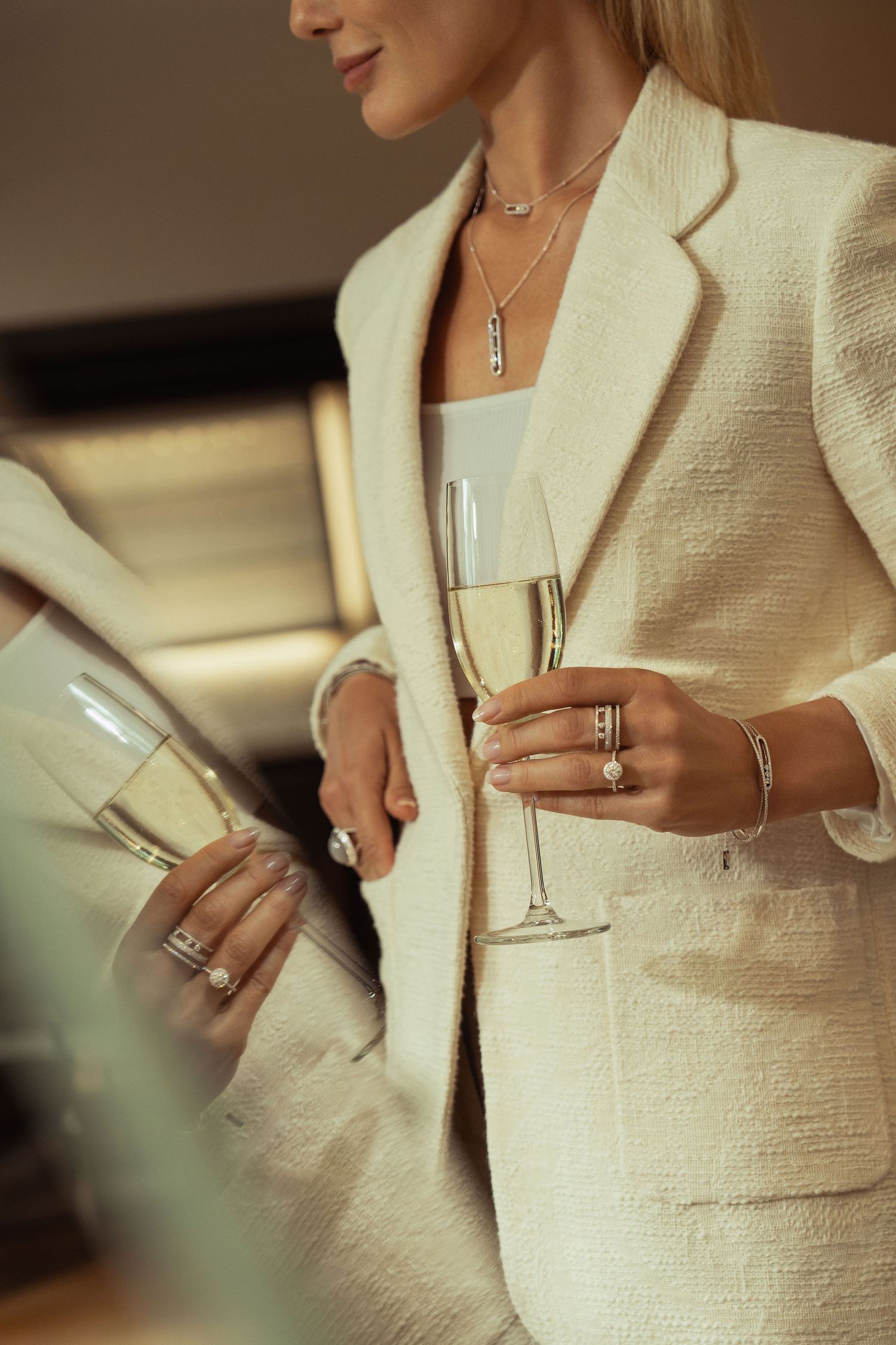 Elegant woman in cream suit holding champagne glass, adorned with diamond rings, bracelets, and layered necklaces.