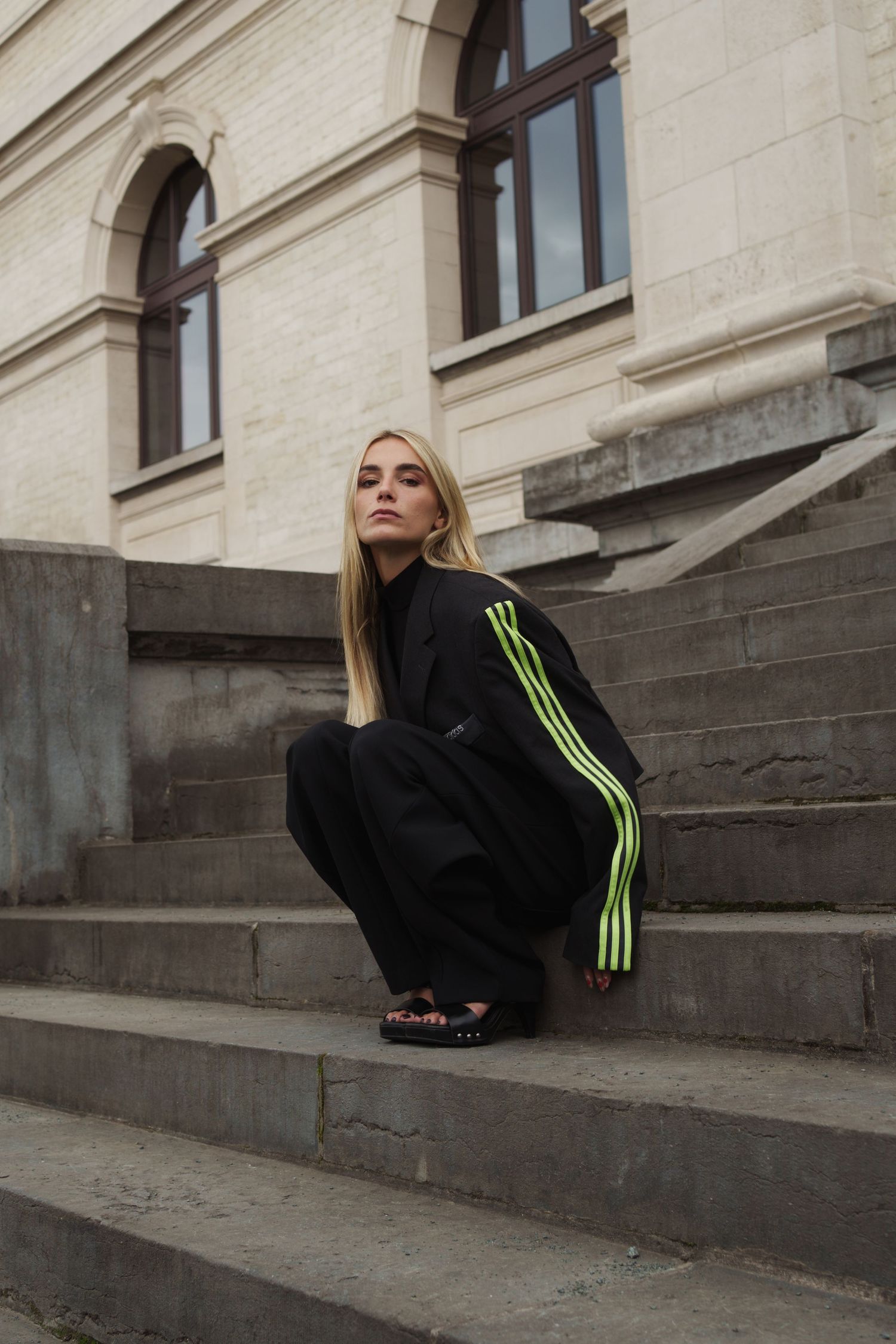Young woman in black Adidas tracksuit with neon green stripes crouching on stone steps outside a classic building.