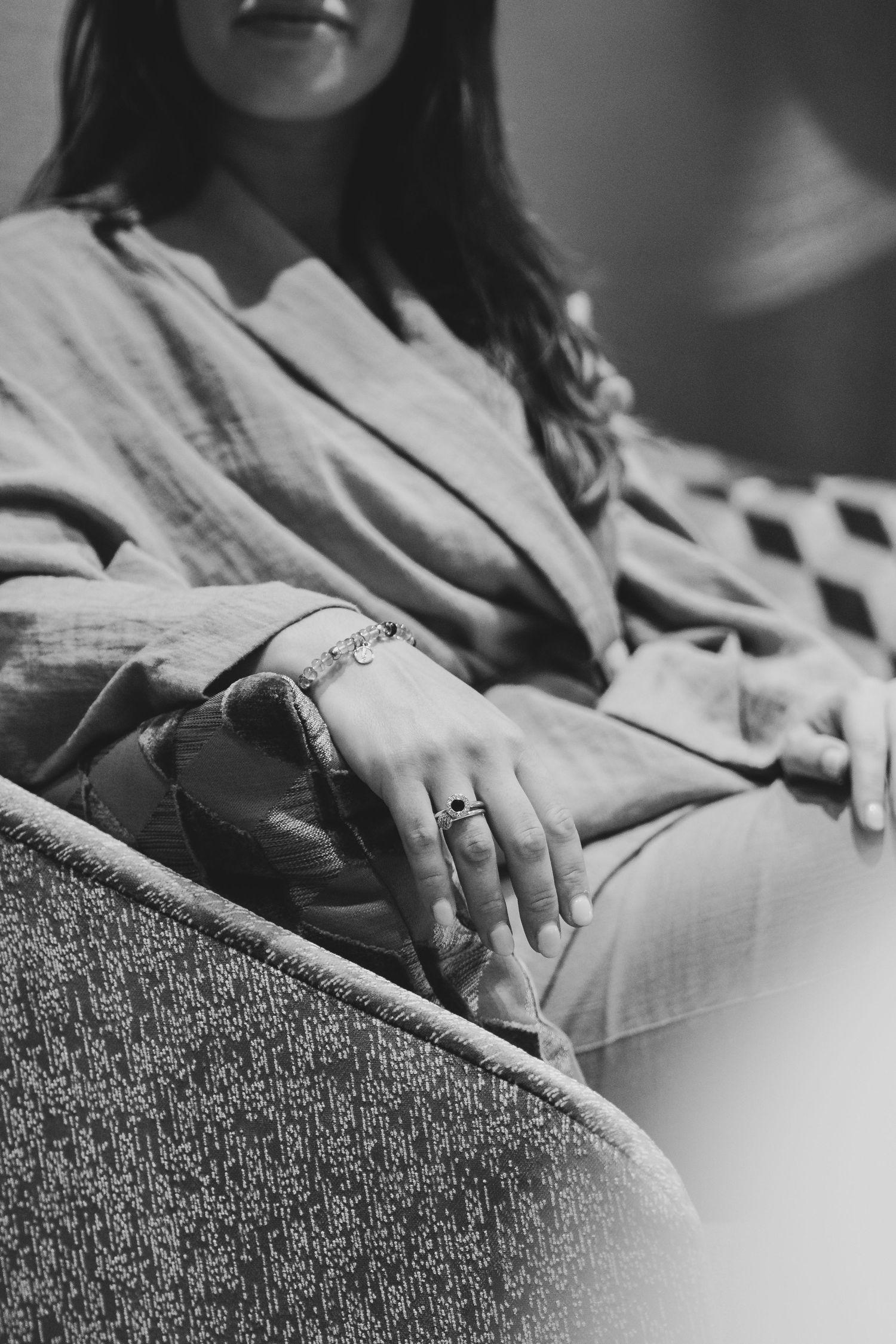 Black and white photo of a woman sitting on a sofa, wearing a bracelet and ring, hands resting on her lap.