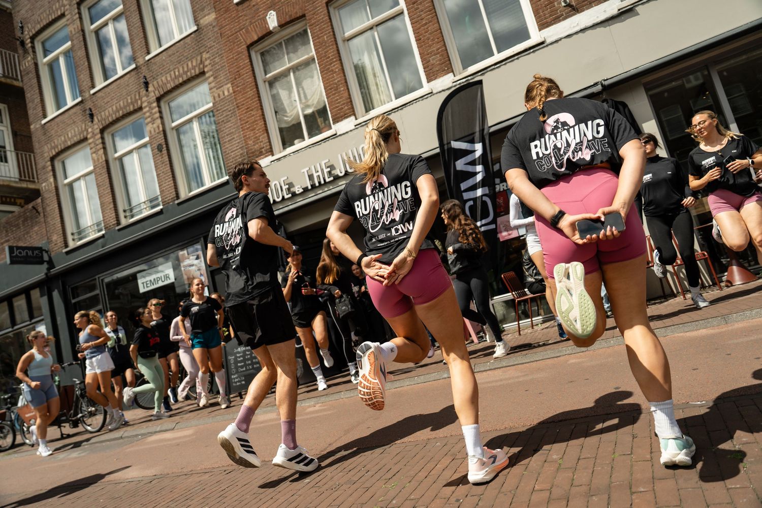 Group of people running on a sunny city street past shops during an outdoor running club event.