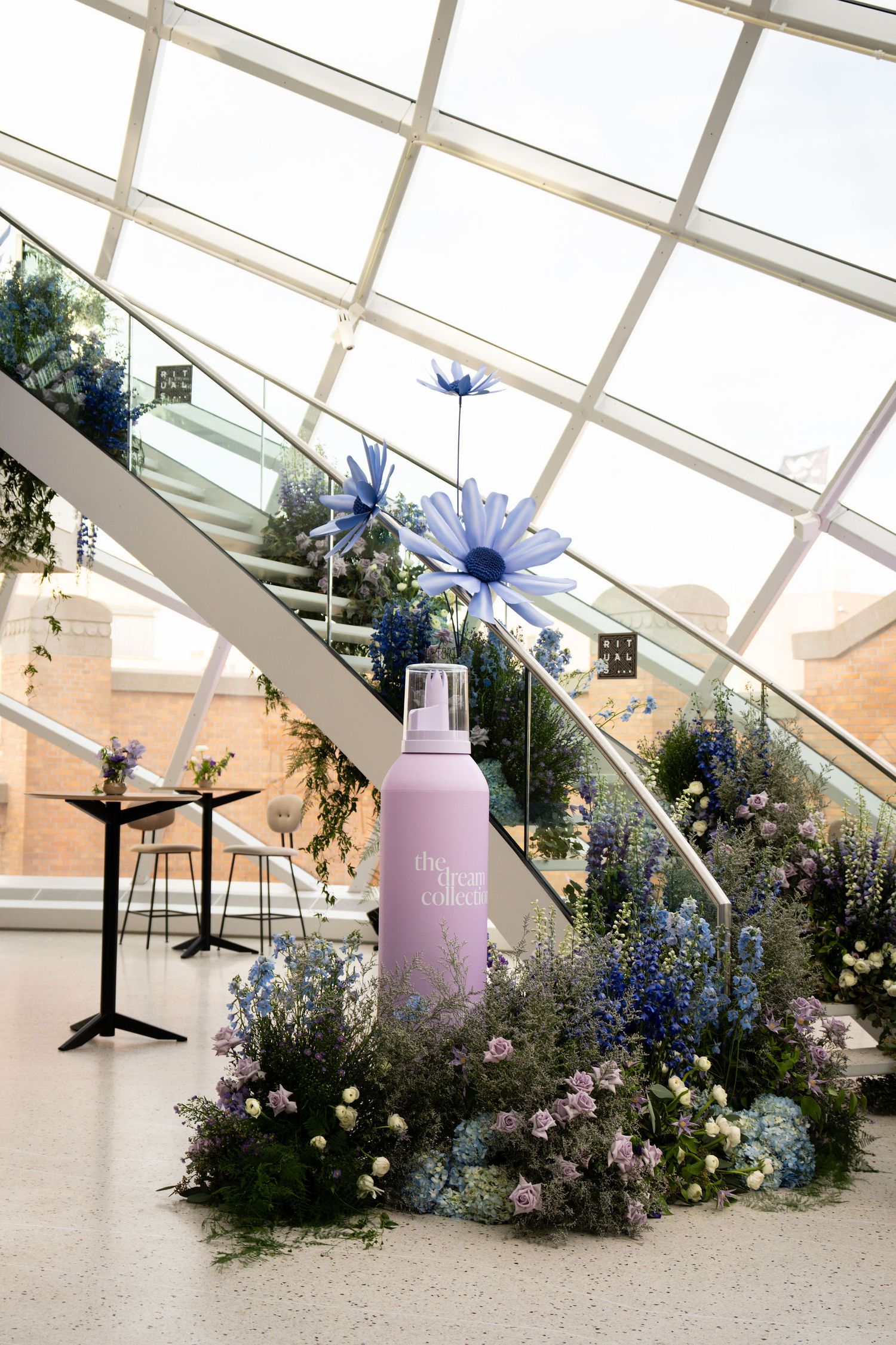 Large pink spray bottle surrounded by blue and purple flowers under a glass ceiling atrium staircase display.