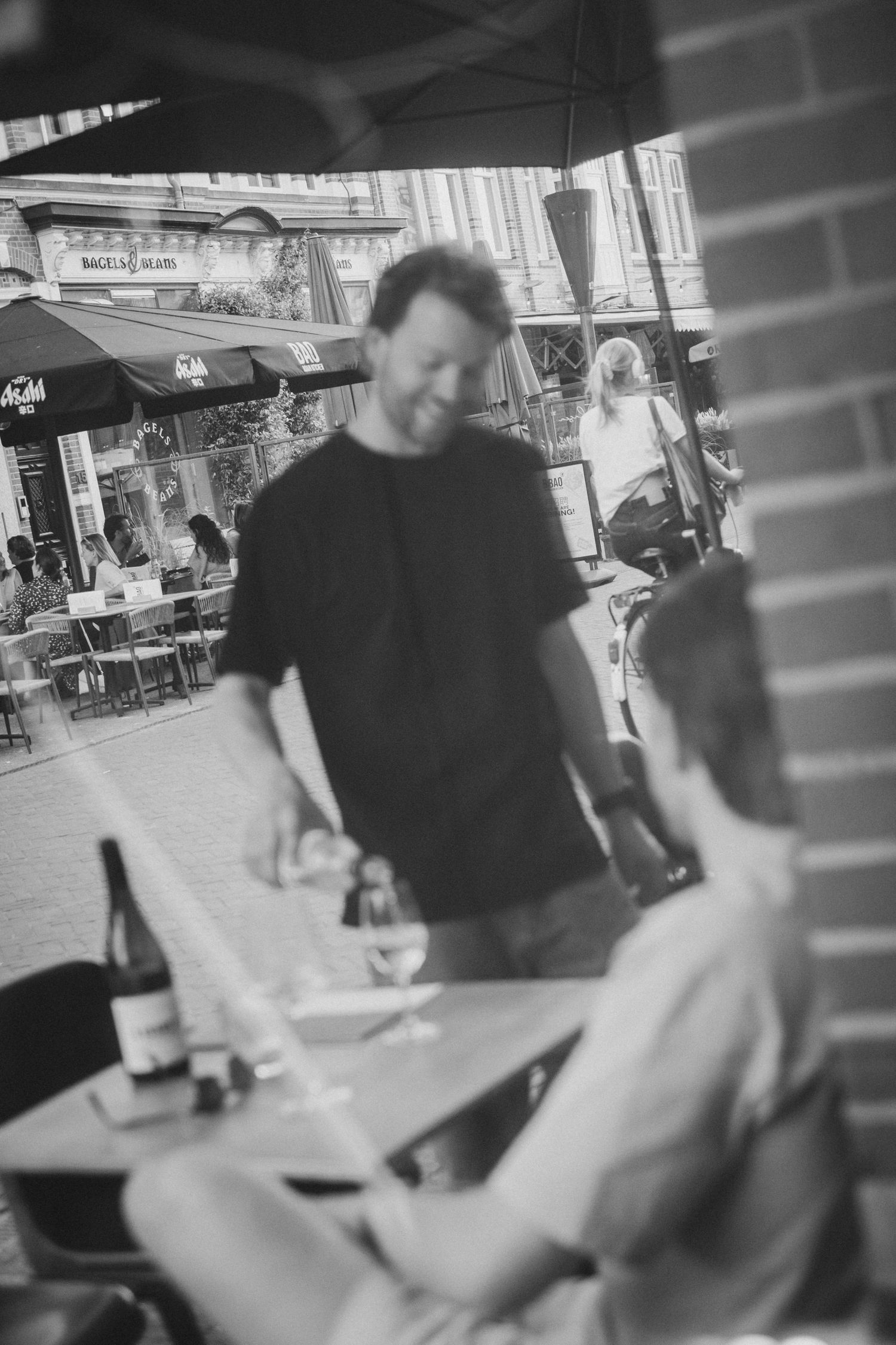 Man in black t-shirt stands at outdoor cafe terrace table in candid black and white photo.