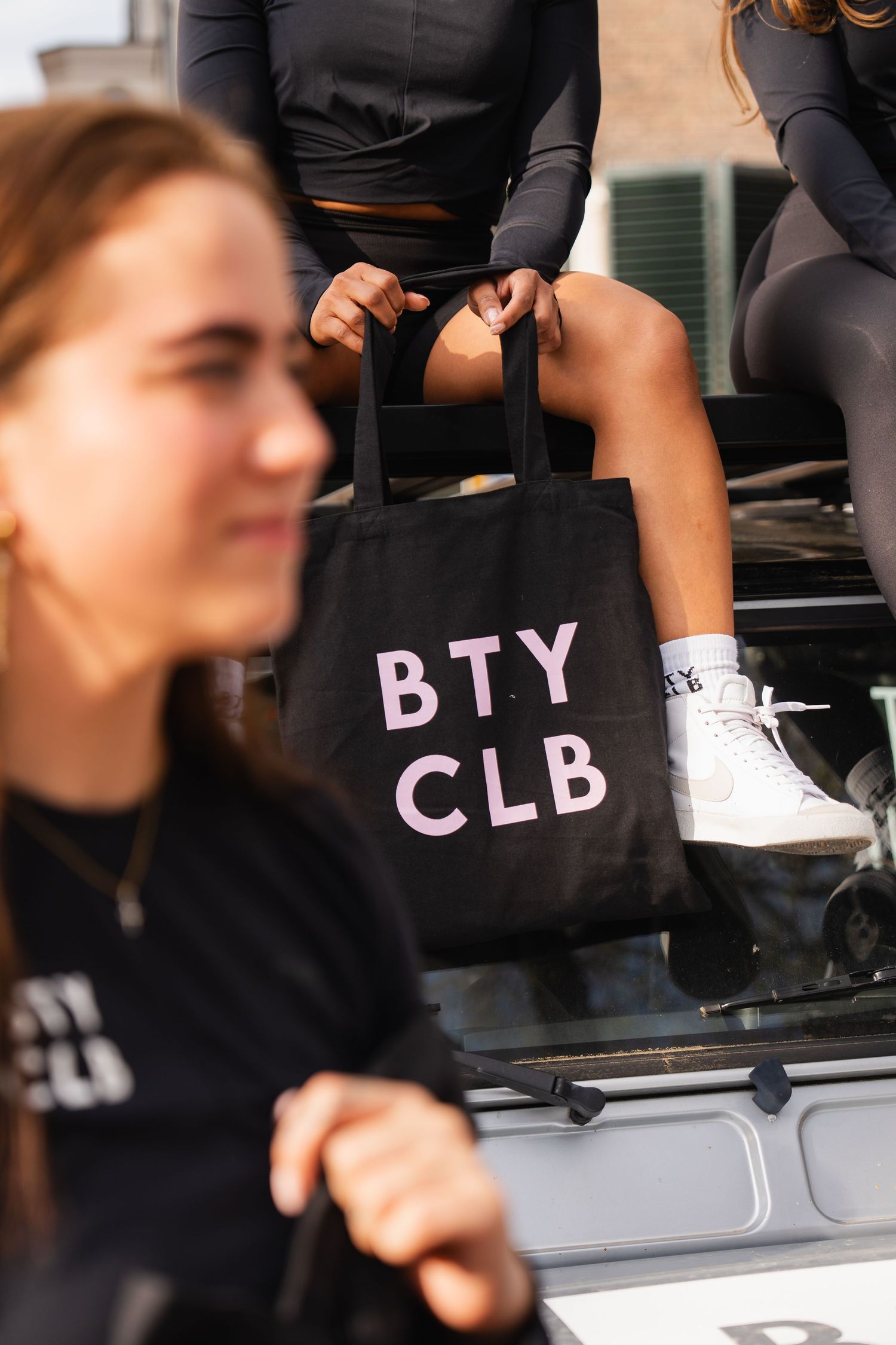 Two women with black BTY CLB tote bag and matching shirts outdoors near a vehicle.