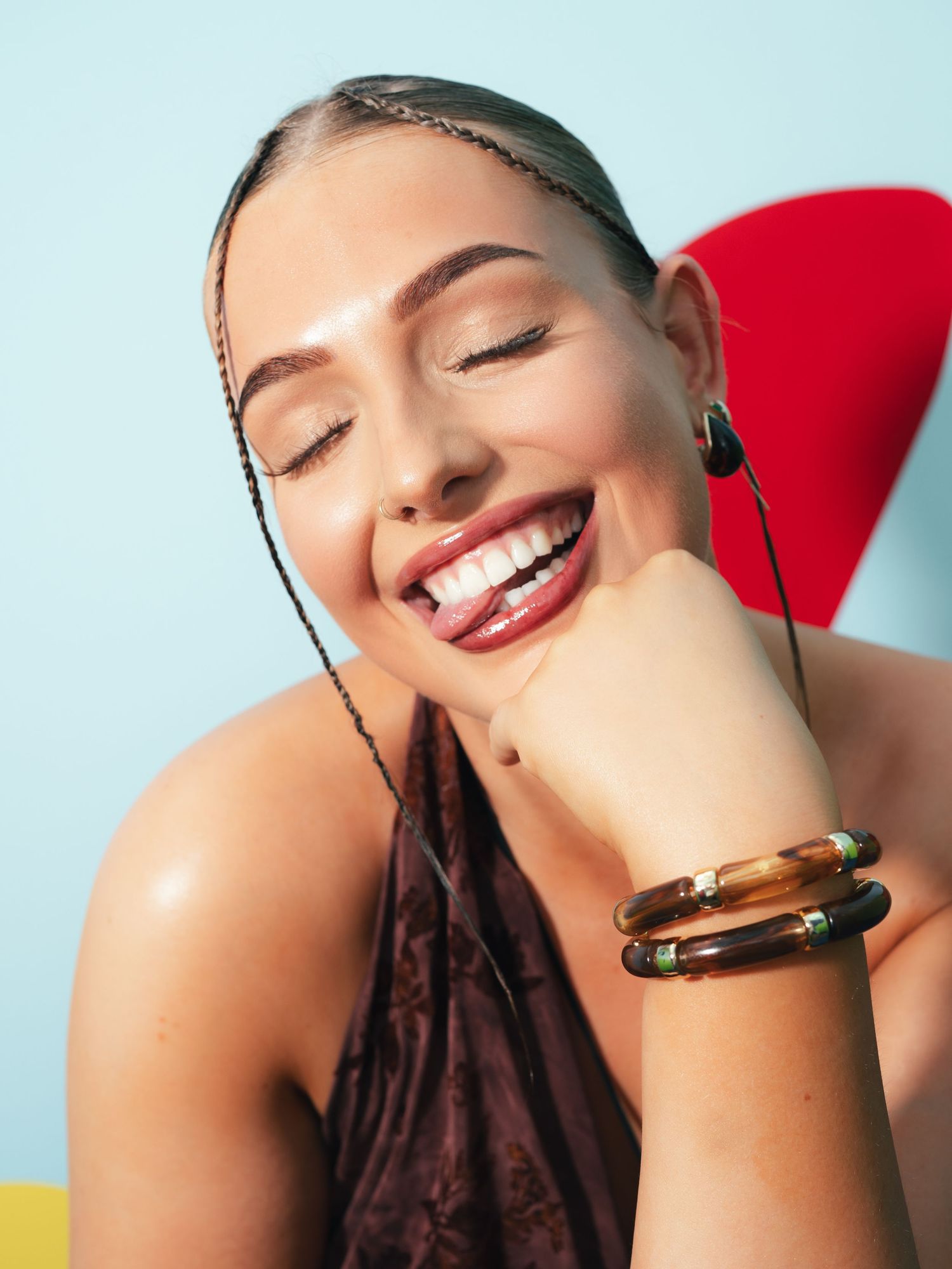 Smiling woman with braids, tortoise shell bracelets, and dark red lips poses against a light blue background.
