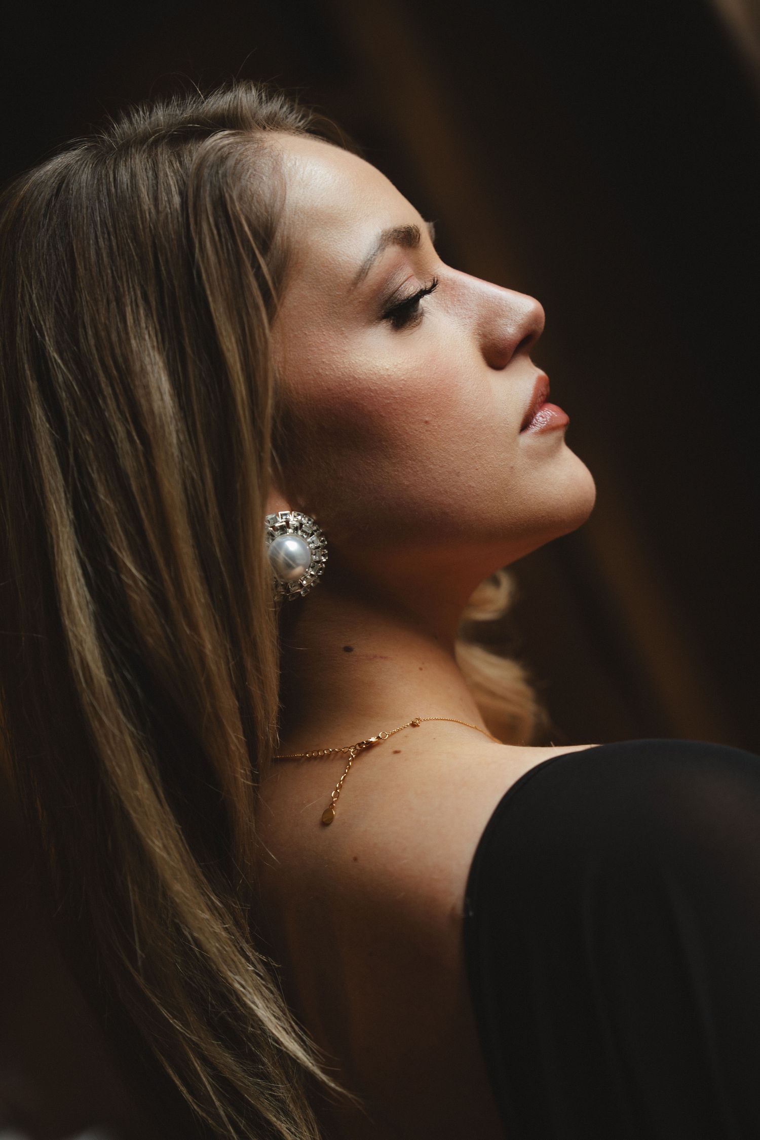 A woman in profile view wearing a large pearl earring and gold necklace, with long blonde hair and a black off-shoulder top.