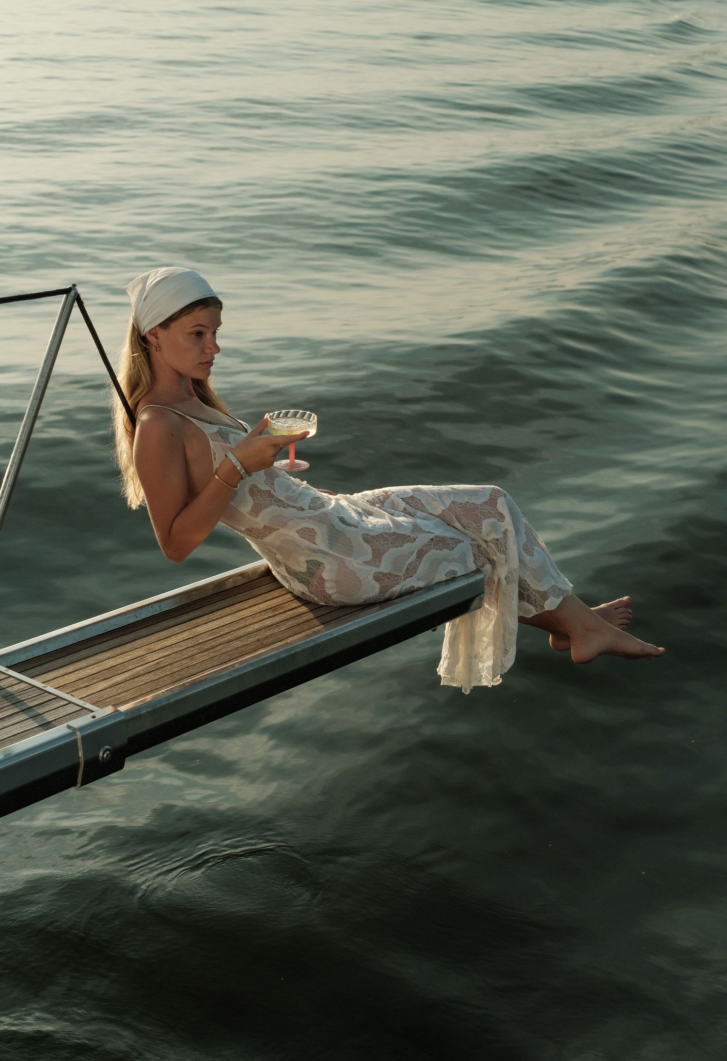 Woman in floral outfit and white headband sitting on a dock over calm water, holding a cocktail glass.