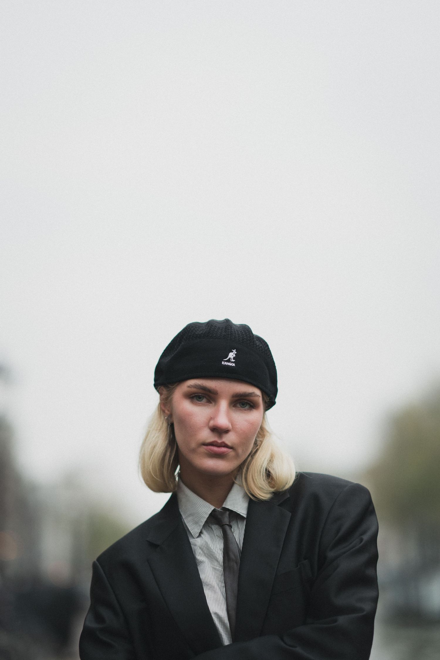 Blonde woman in black Kangol beret and oversized blazer poses outdoors with serious expression.