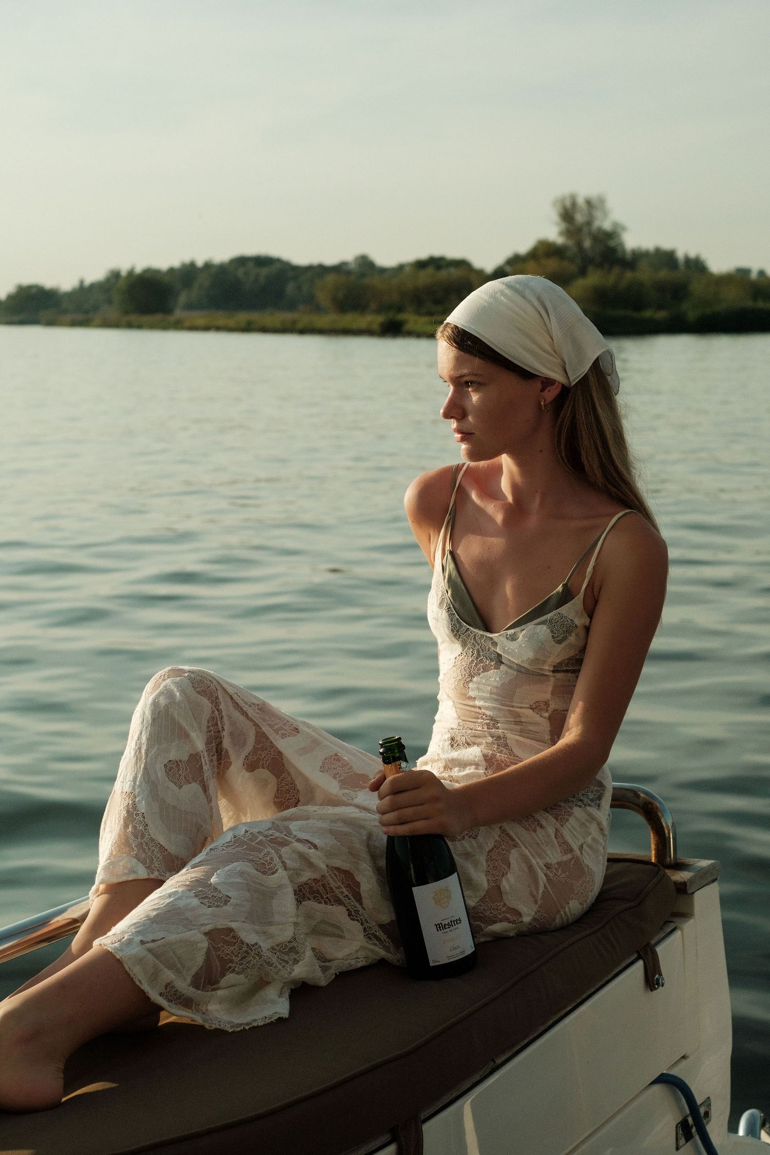 Woman in lace dress holding wine bottle seated on boat bow with serene lake and green trees in background.