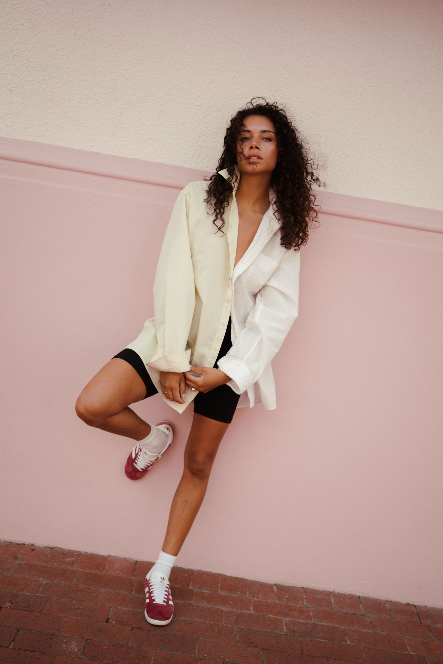 Young woman in white blazer, black bike shorts, and burgundy Adidas sneakers posing against a pink wall.
