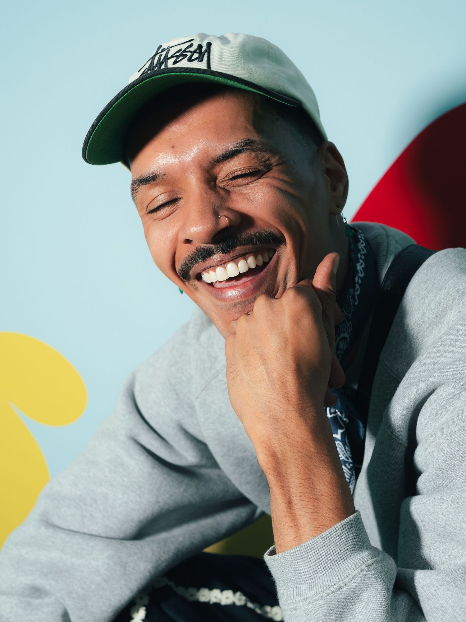 Smiling man wearing a Stussy cap and gray sweatshirt laughing joyfully against a colorful blue background.