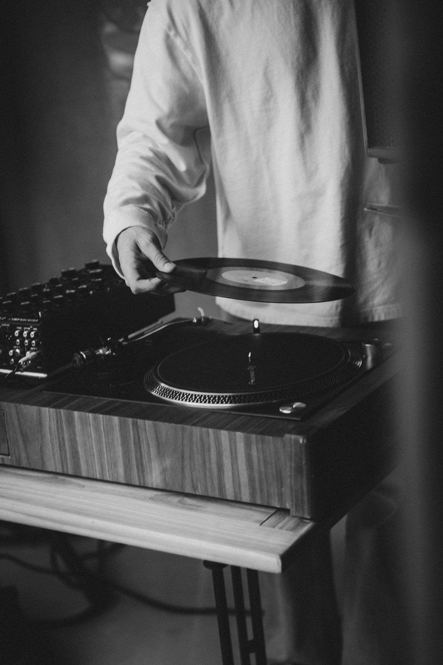 Black and white photo of a DJ scratching a vinyl record on a turntable with a mixer at an indoor event.