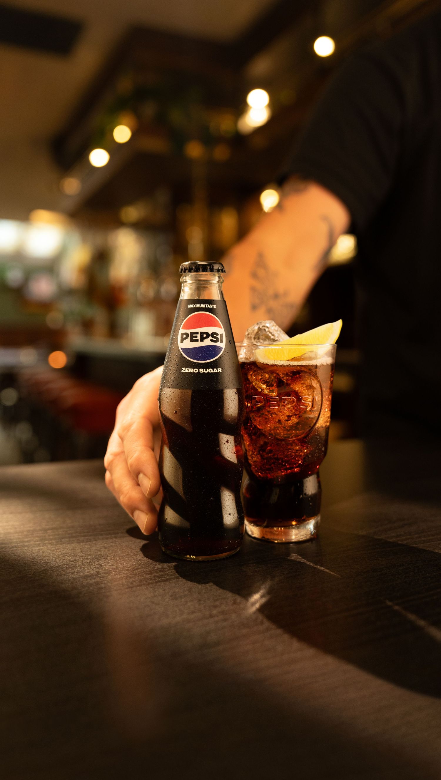Tattooed hand holding a Pepsi Zero Sugar bottle beside a cola glass with ice and lemon slice on a dark bar counter.