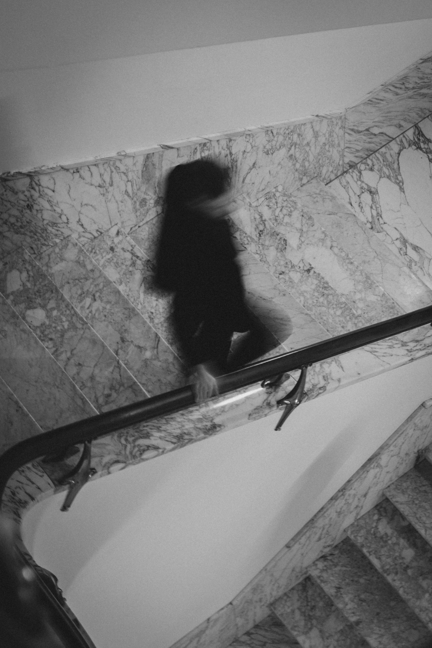 Blurred black and white photo of a person descending marble stairs with a dark handrail, creating a ghostly motion effect.