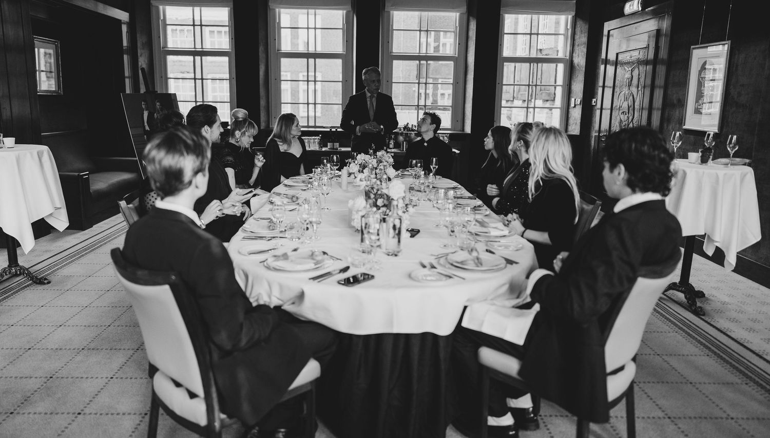 Black and white photo of elegantly dressed guests seated at a round dining table in a formal wood-paneled banquet room.