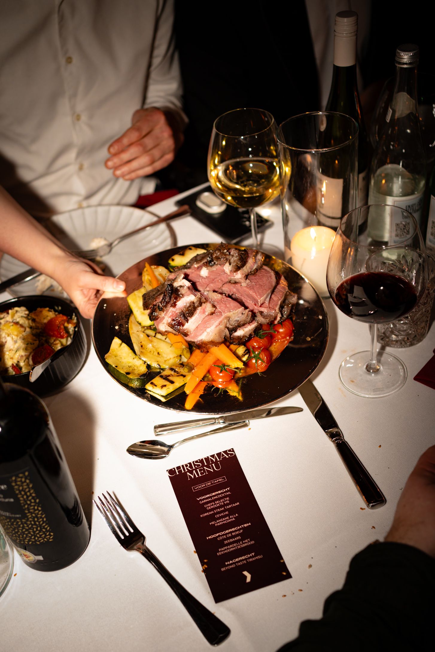 Elegant Christmas dinner table with sliced roasted meat, grilled vegetables, wine glasses, and a festive holiday menu card.