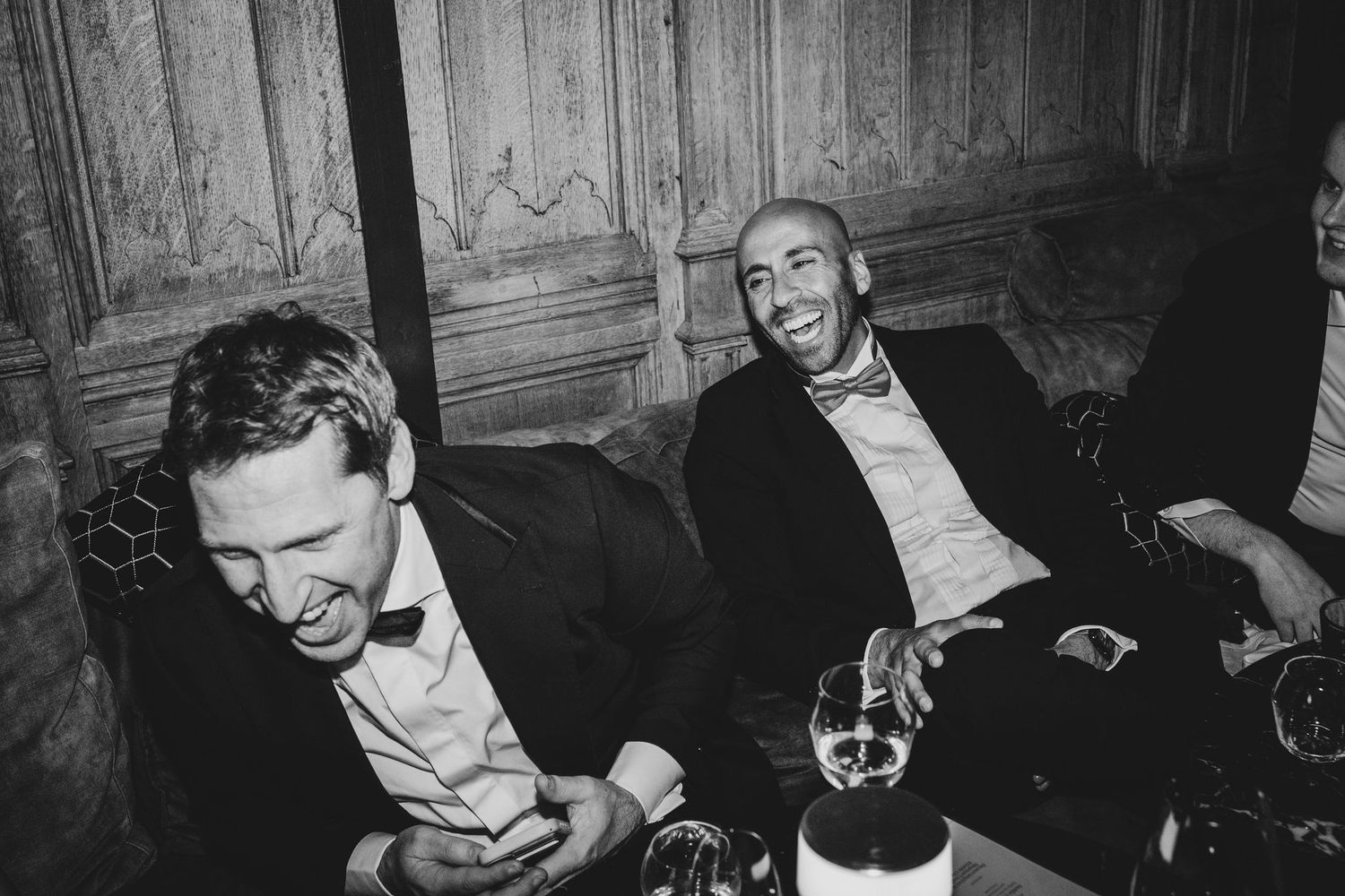 Two men in tuxedos laughing at a formal event, black and white candid photography.