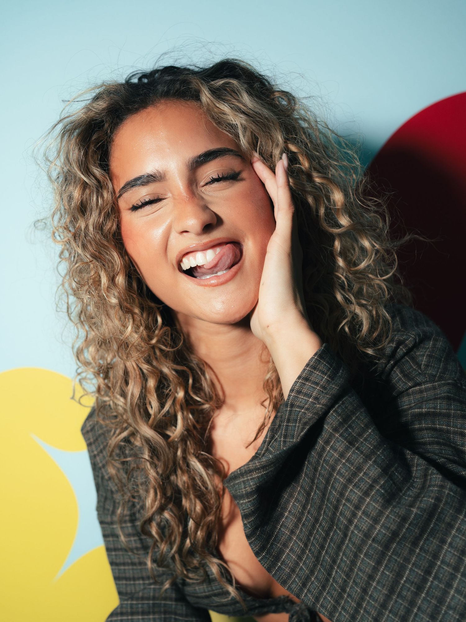 Playful woman with curly blonde hair sticking out tongue, hand on cheek, wearing a plaid blazer against a colorful backdrop.