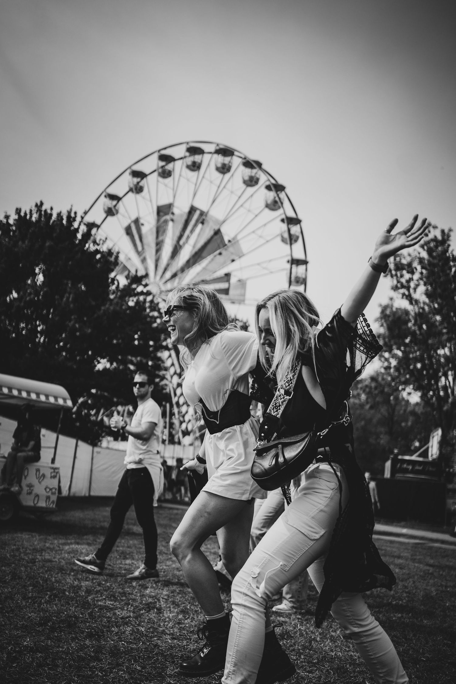 Two women dancing joyfully at an outdoor festival with a Ferris wheel in the black and white background.