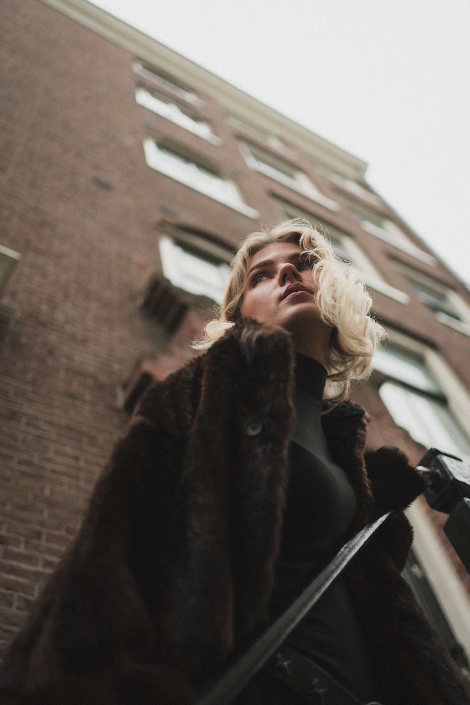 Blonde woman in fur coat gazing upward beside brick building, shot from low angle perspective.