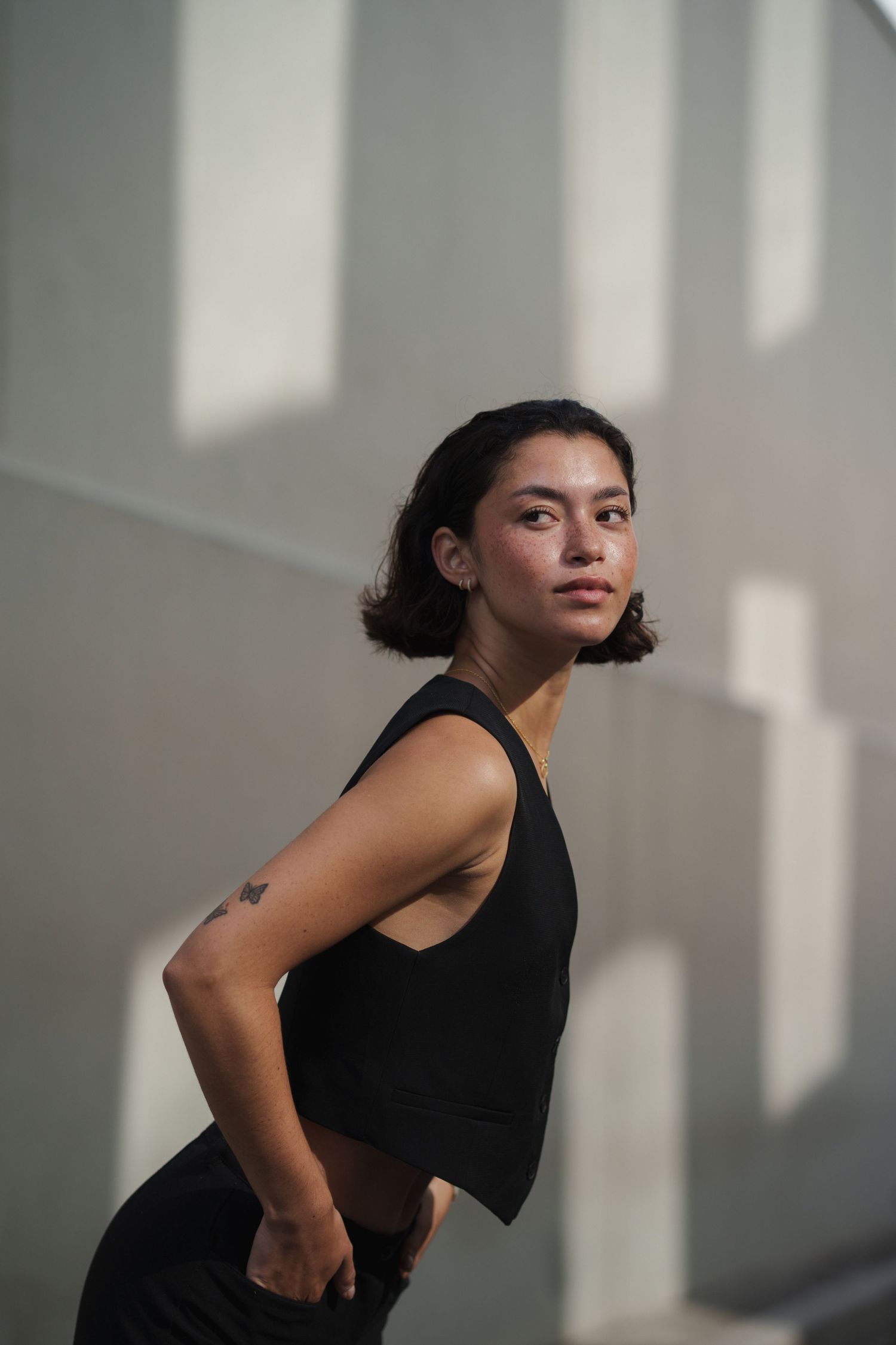 Young woman in black crop top and pants poses confidently against a gray wall with soft shadows.