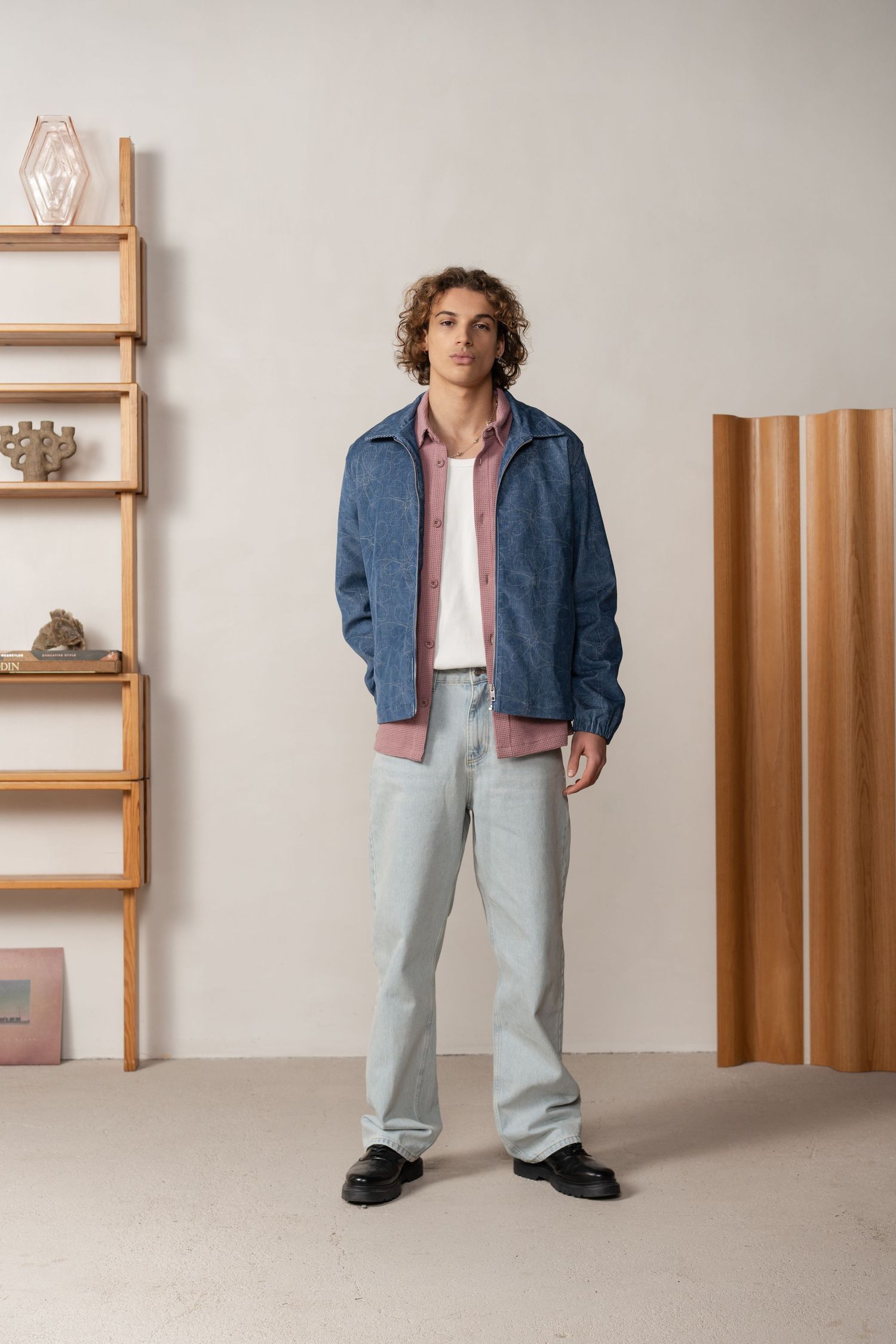 Man in blue jacket, pink shirt, and light jeans stands in a minimalist room with wooden shelving.