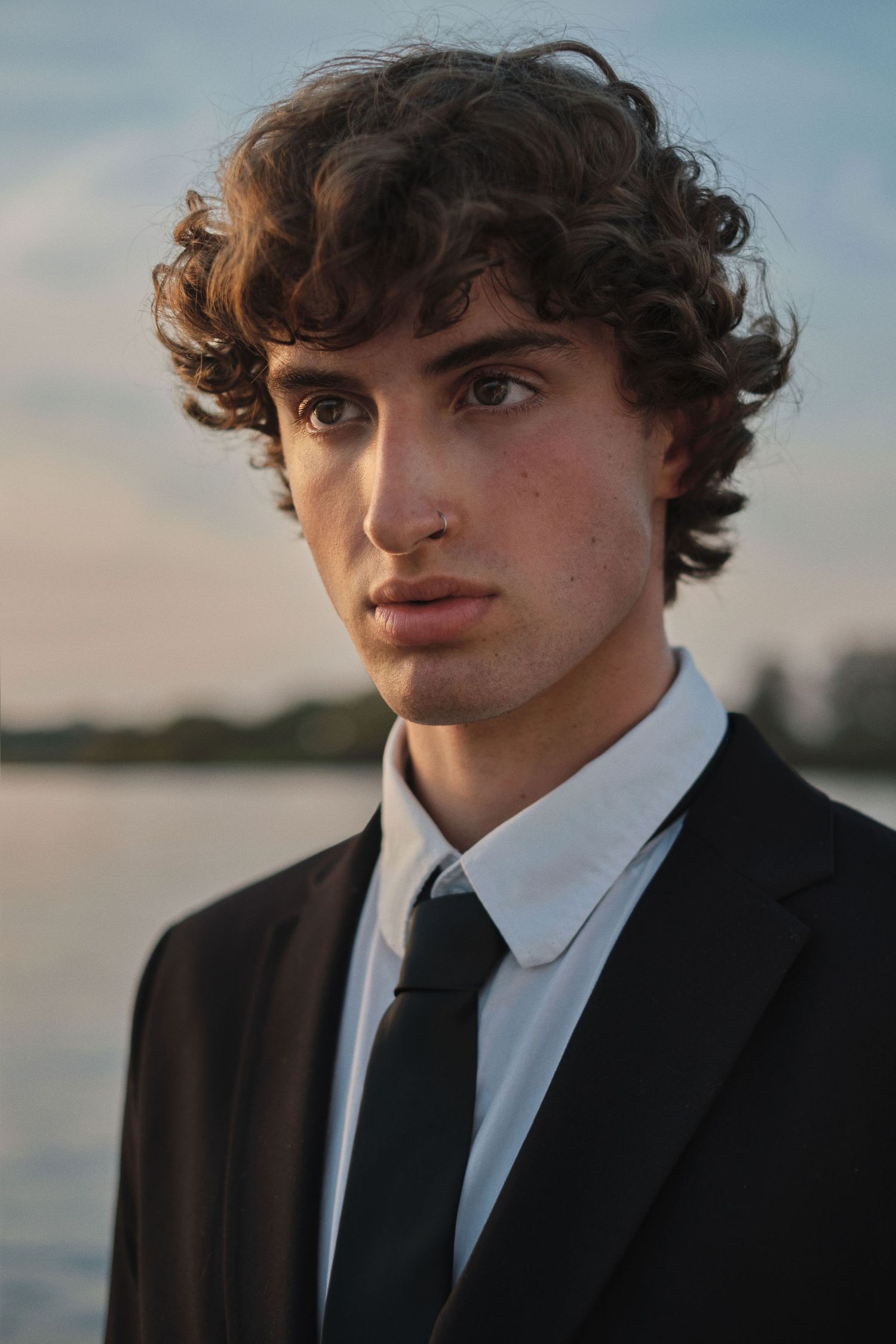 Young man with curly hair in black suit and tie poses outdoors near water at dusk.