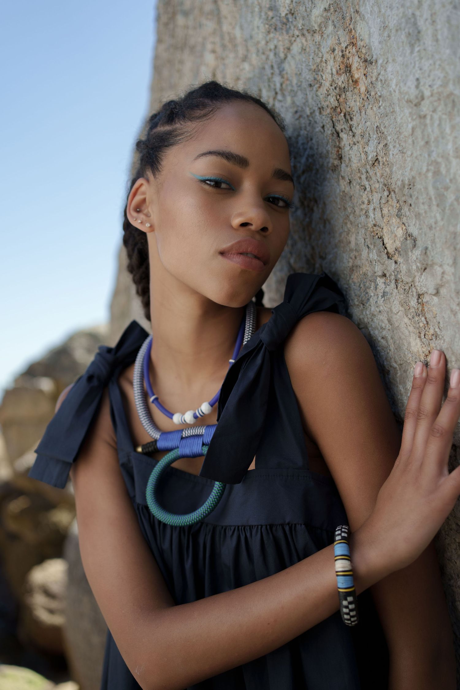 Young Black woman in black dress with colorful beaded necklaces and bracelet leaning against a rock outdoors.
