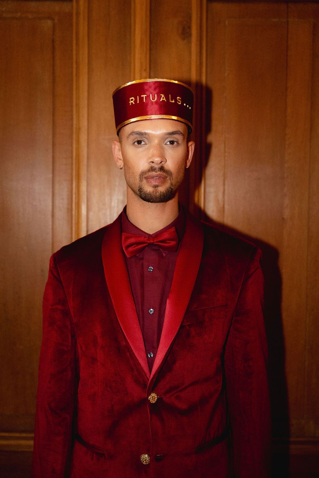 Man in red velvet suit and bow tie wearing a Rituals branded bellhop hat against wood paneling.
