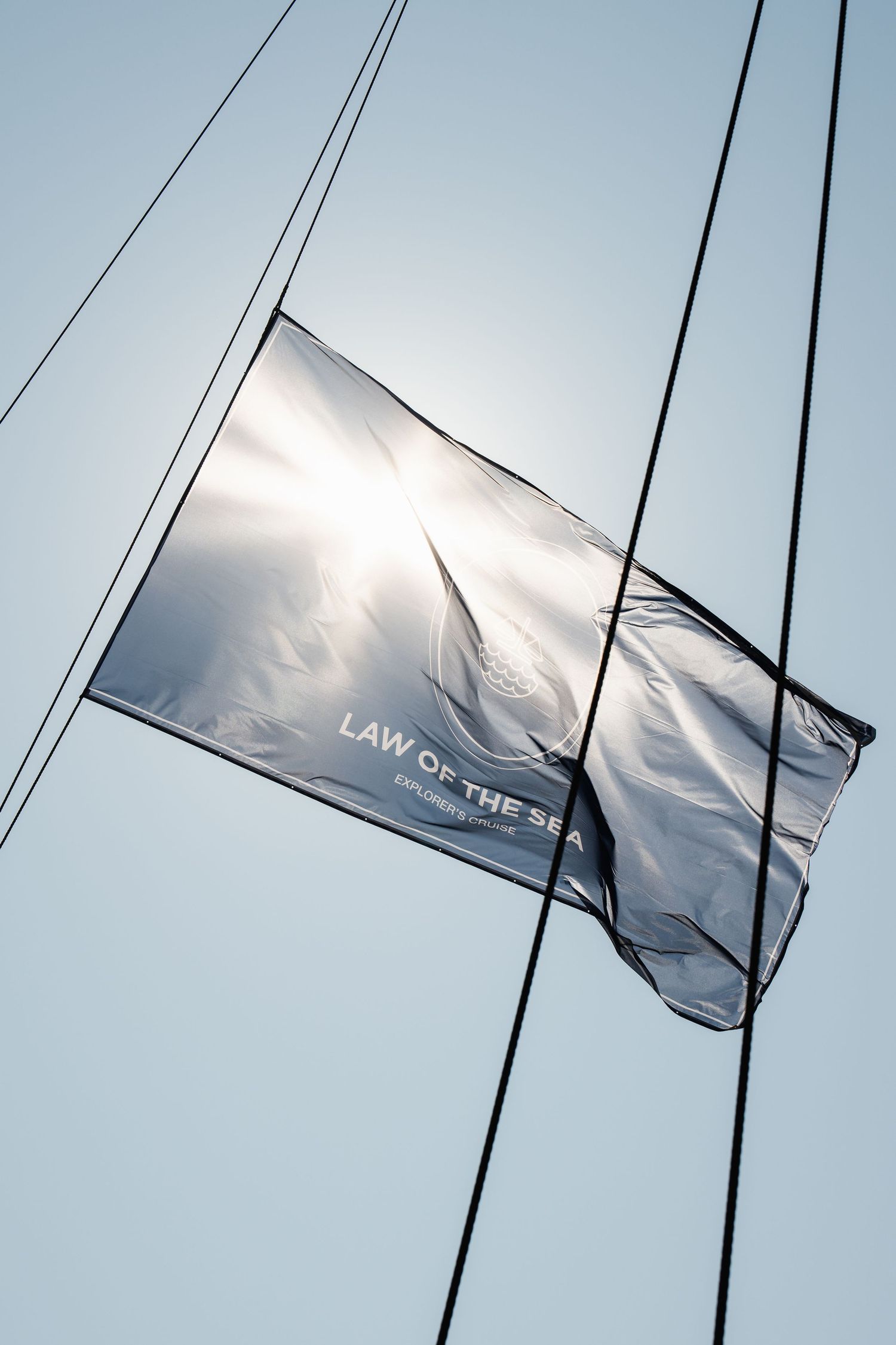 A silver Law of the Sea flag backlit by sunlight, hanging from rigging against a clear blue sky.