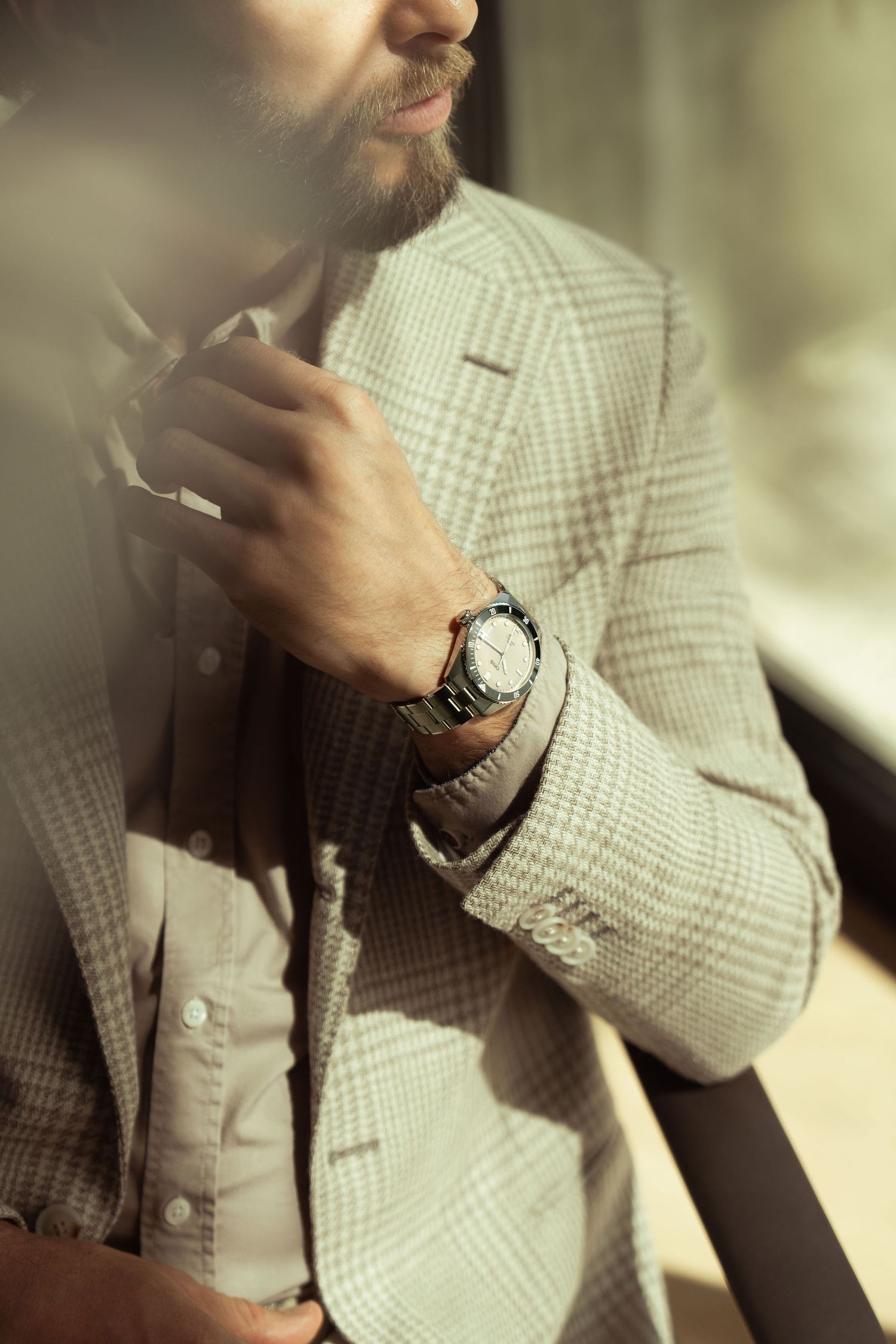 Bearded man in plaid blazer adjusting collar, wearing silver watch, bathed in warm natural light.