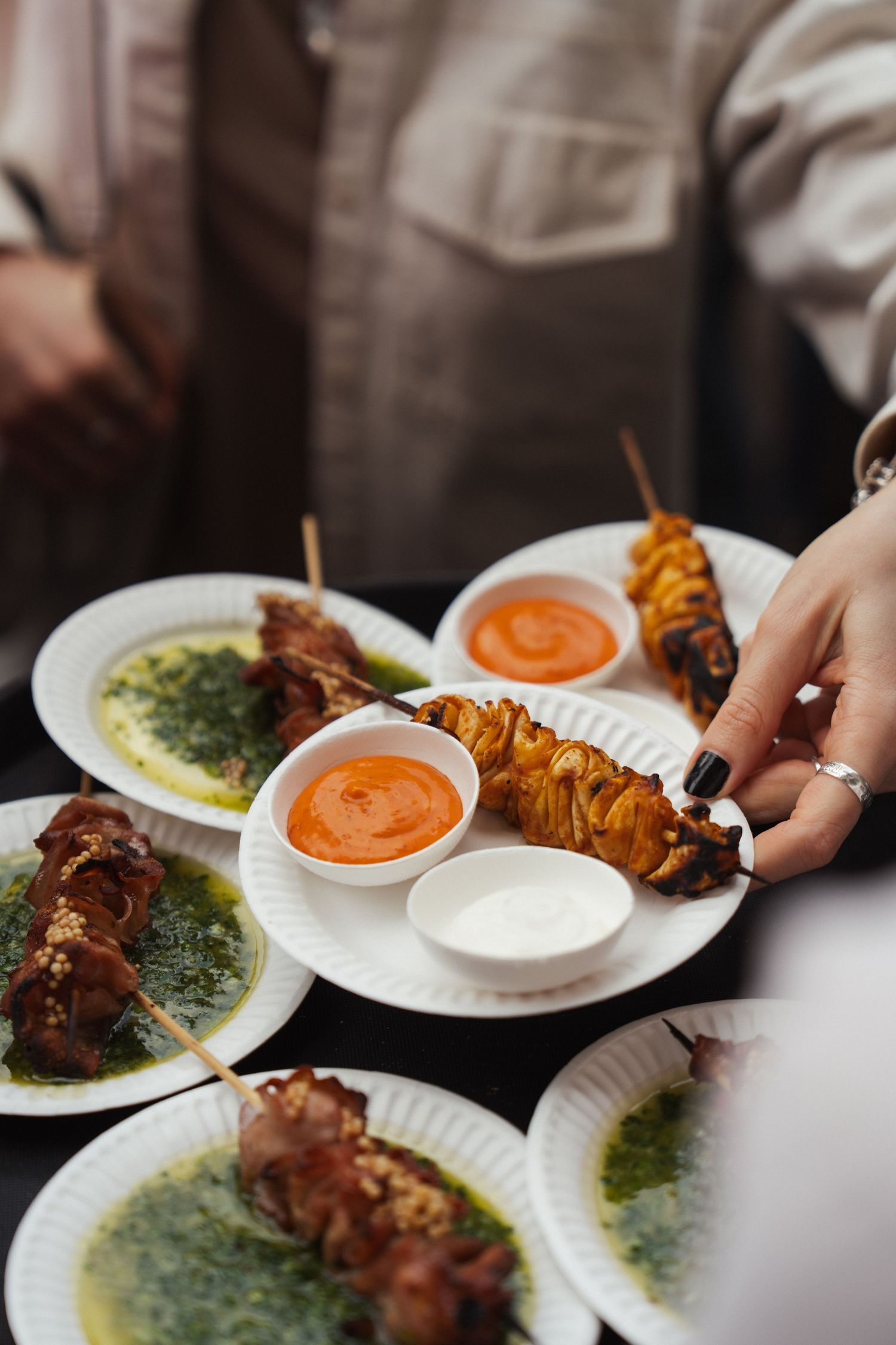 Grilled meat skewers on white plates with orange and white dipping sauces, garnished with herb oil.