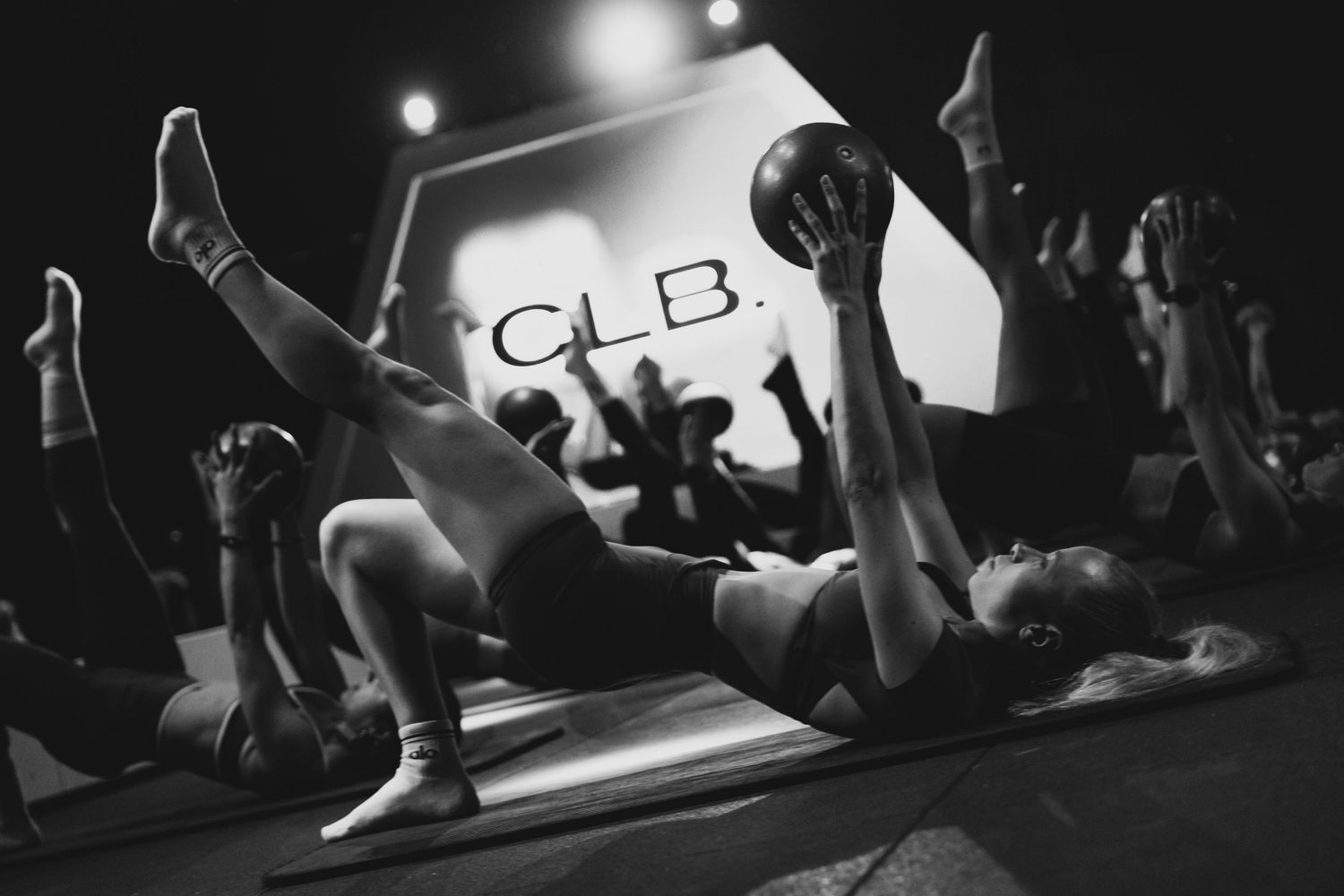 Black and white photo of women doing Pilates exercises with medicine balls in a dark studio with CLB branding.