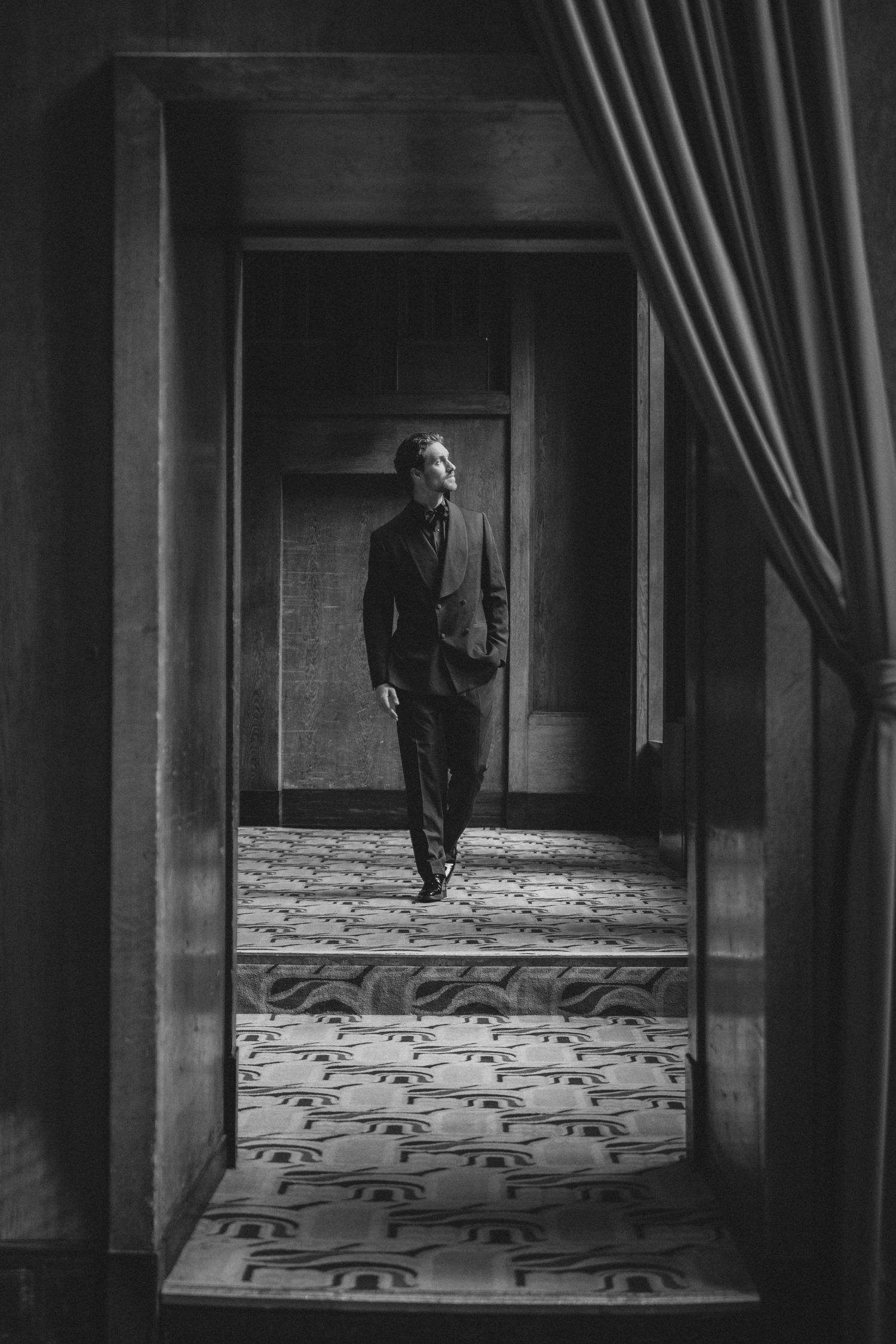 Black and white photo of a man in a suit walking through an elegant hallway with patterned carpet and draped curtains.