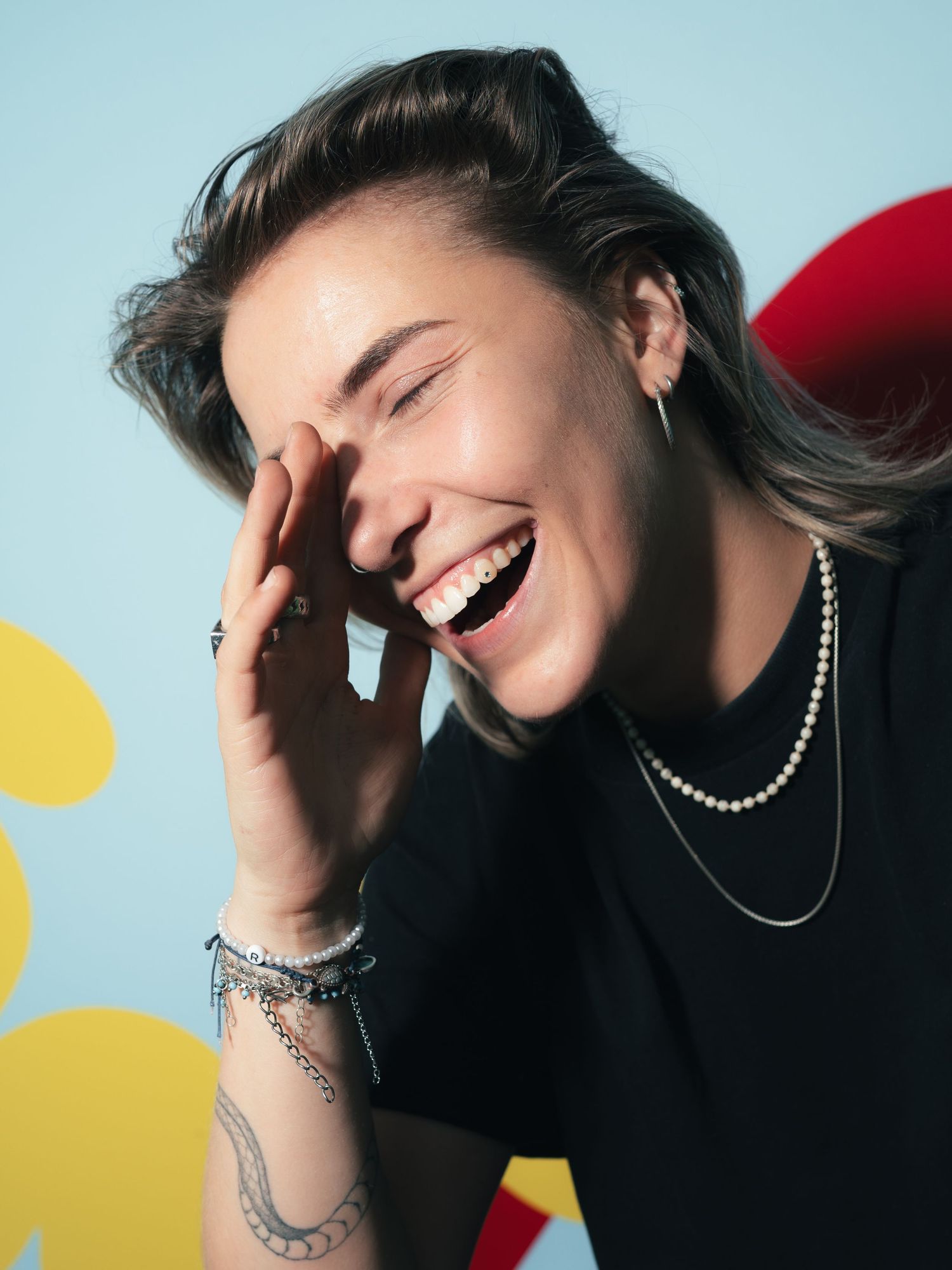Laughing young woman with tattoos, pearl necklace, and bracelets poses against a light blue background.