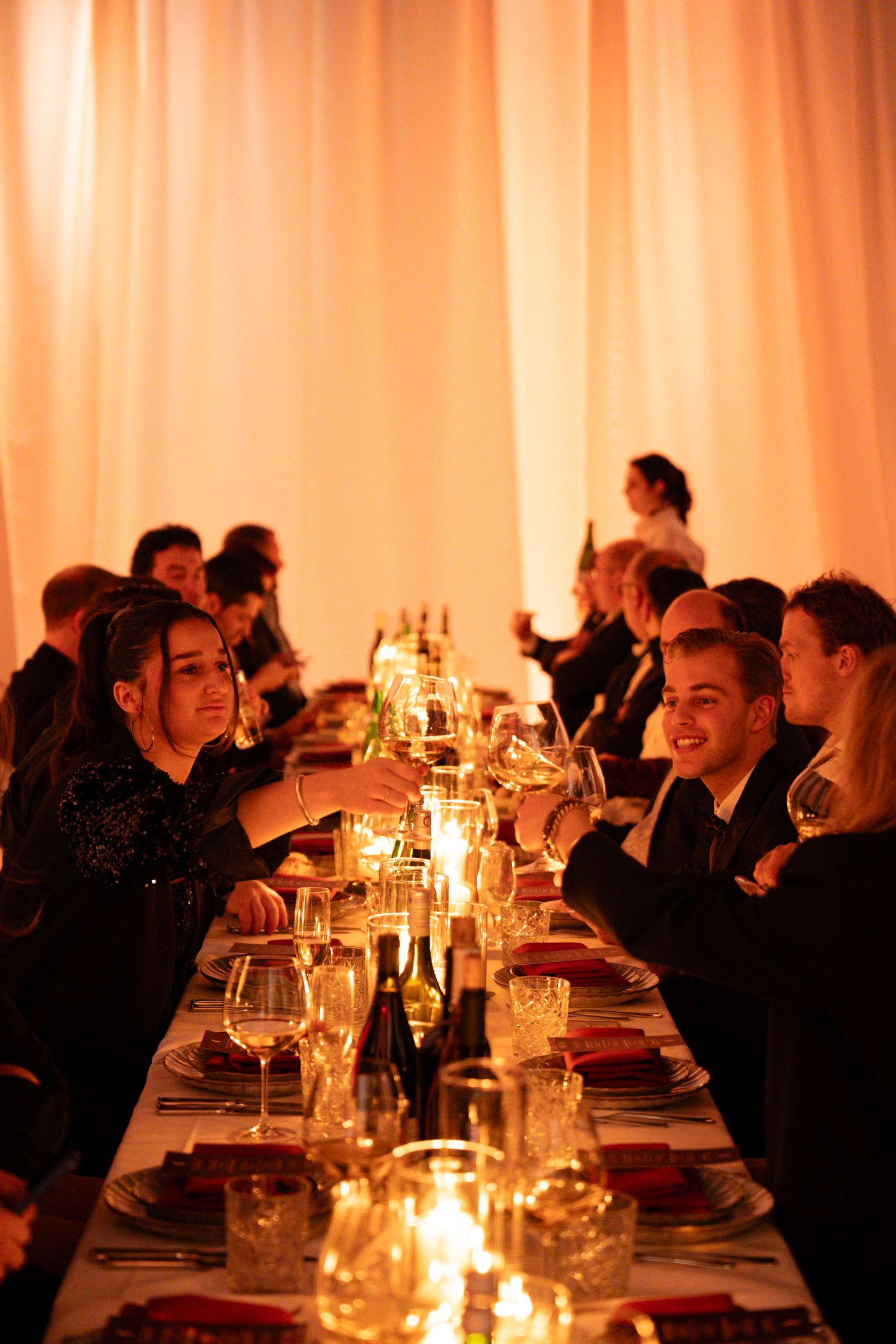 Guests enjoy a candlelit dinner party at a long table with warm amber lighting and wine glasses raised in a toast.