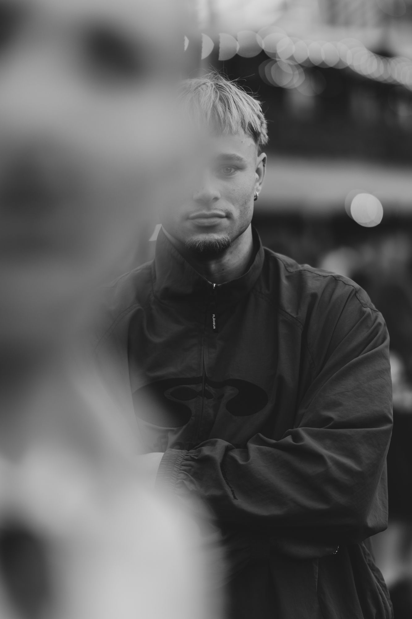 Black and white portrait of a young man with blonde hair wearing a dark jacket, arms crossed, with blurred bokeh background.