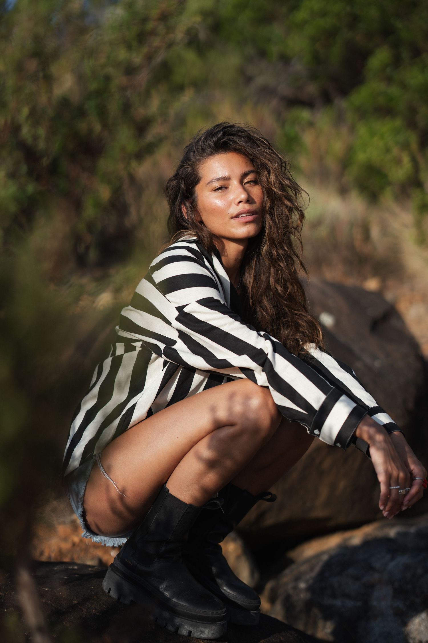 Woman in striped shirt and boots crouching on rocks outdoors in natural sunlight with green foliage background.