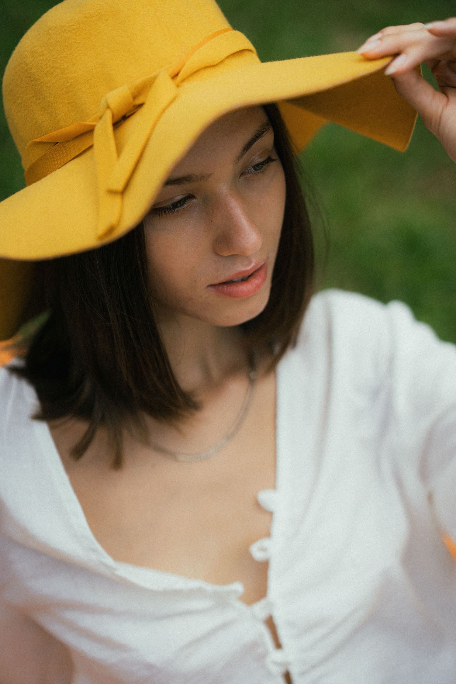 Young woman in white blouse wearing a wide-brim yellow sun hat outdoors, looking down gracefully.