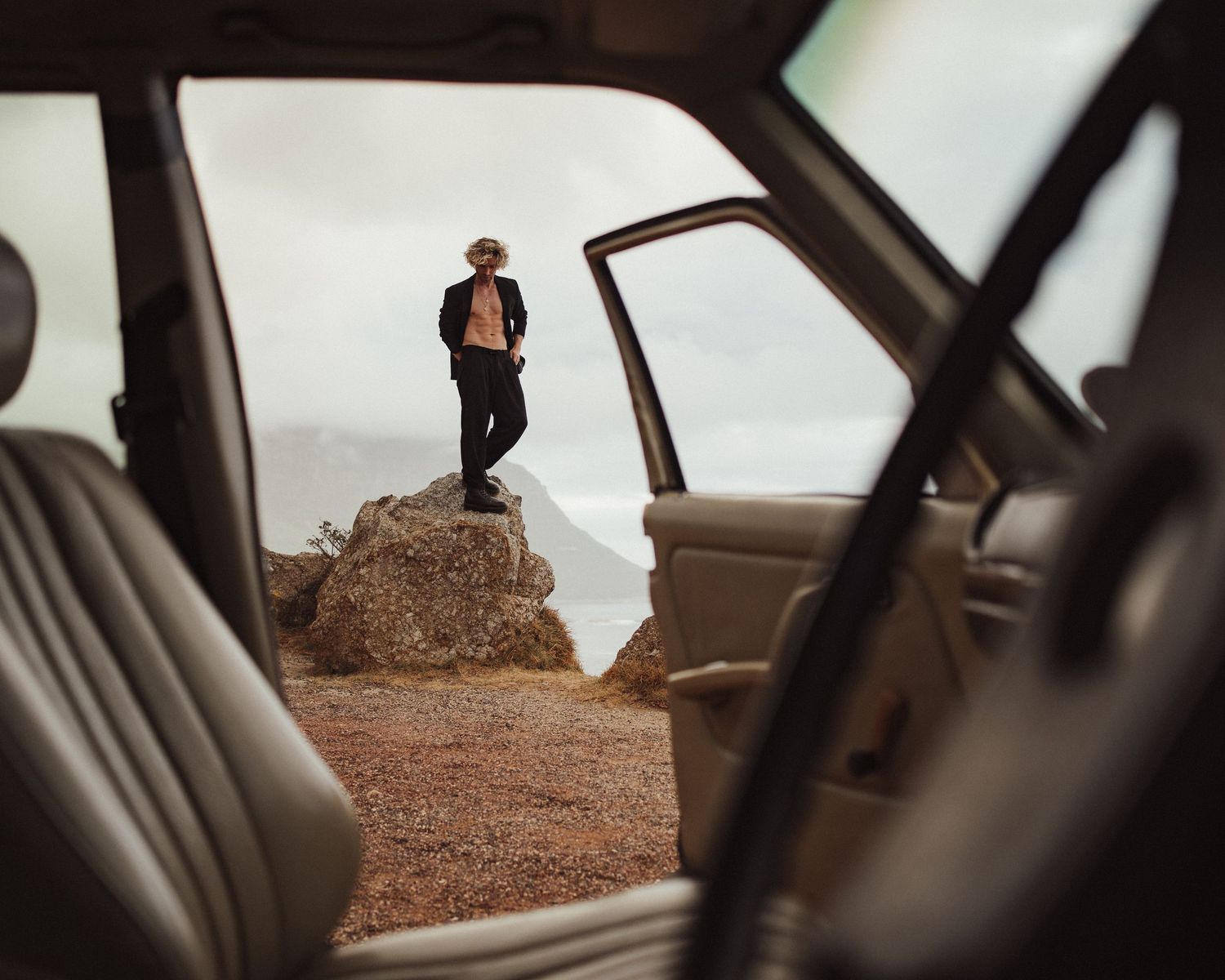 Man standing on a rocky coastal overlook viewed through an open vintage SUV door on a cloudy day.