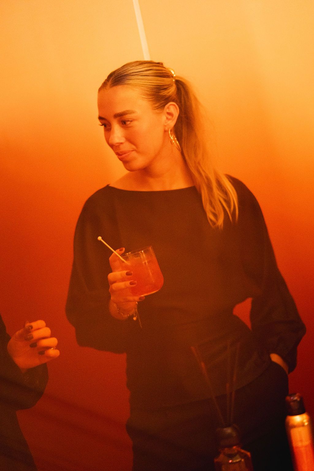 Young woman in black sweater holding a cocktail with a straw at a party under warm orange lighting.