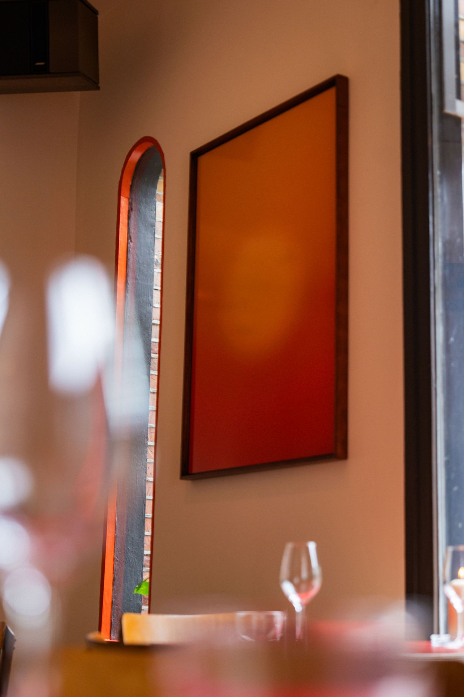 Warm restaurant interior with red-orange framed artwork on beige wall, arched mirror, and wine glasses on table.