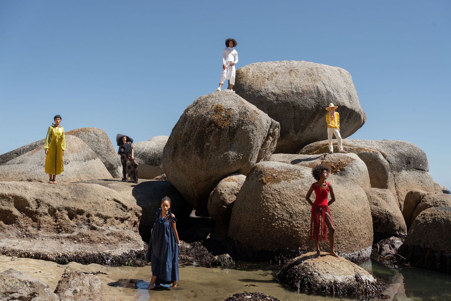 People in colorful outfits posed on large coastal boulders under a clear blue sky.