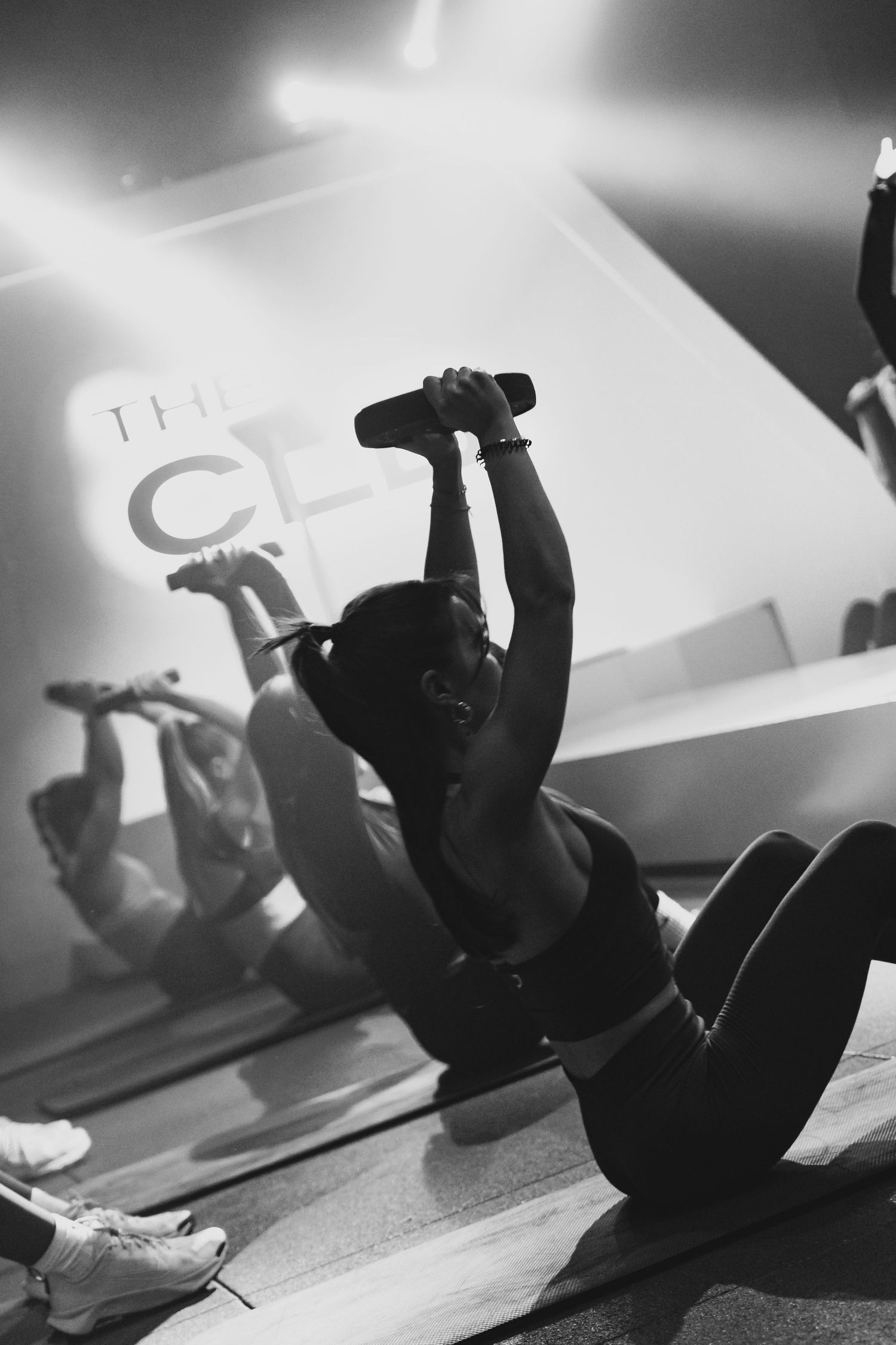 Woman performing sit-ups with a dumbbell overhead in a dark gym class, black and white fitness photo.