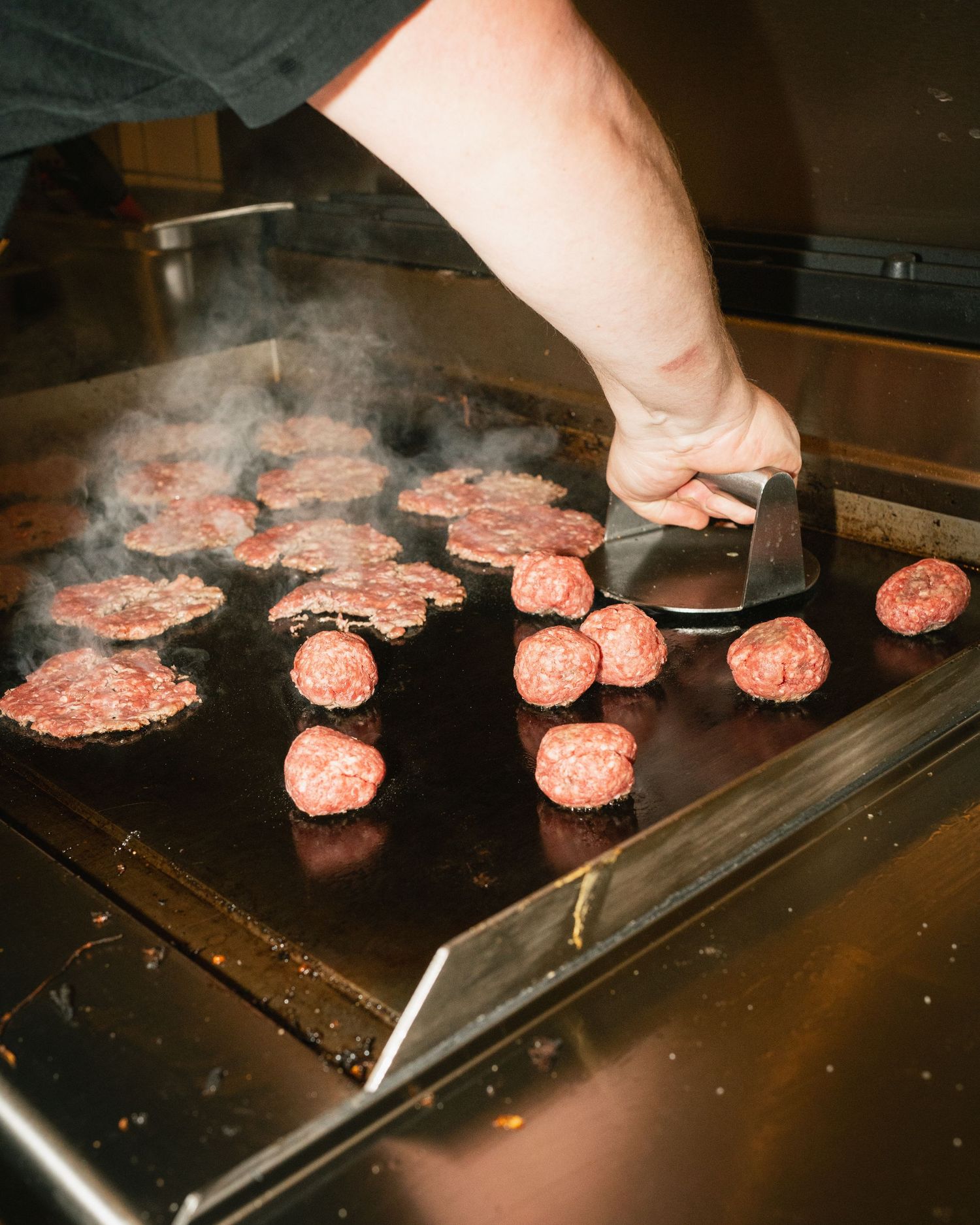 Chef smashing burger patties on a hot commercial griddle with steam rising in a restaurant kitchen.
