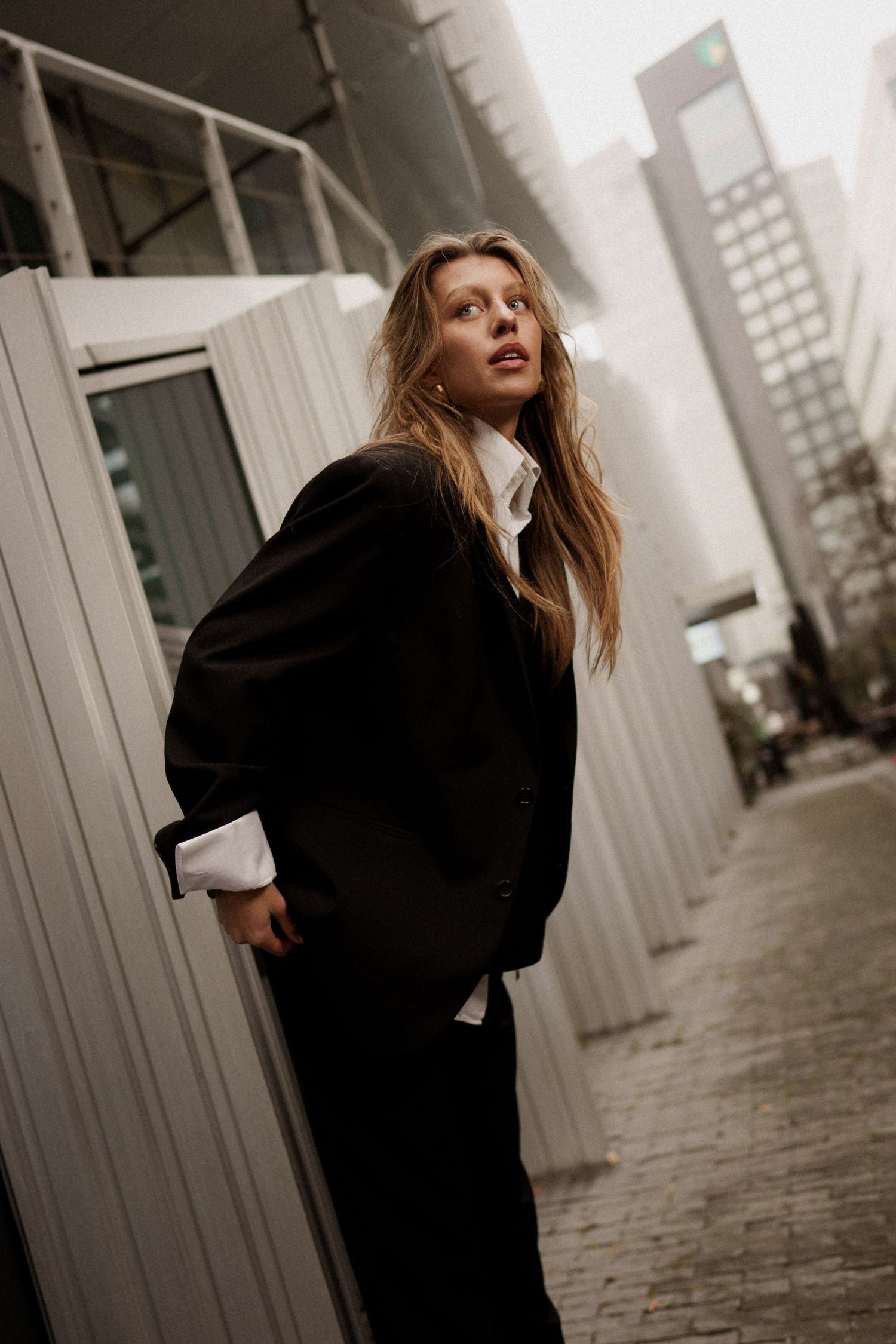 Woman in oversized black blazer and white shirt poses on urban cobblestone street with modern skyscrapers in background.