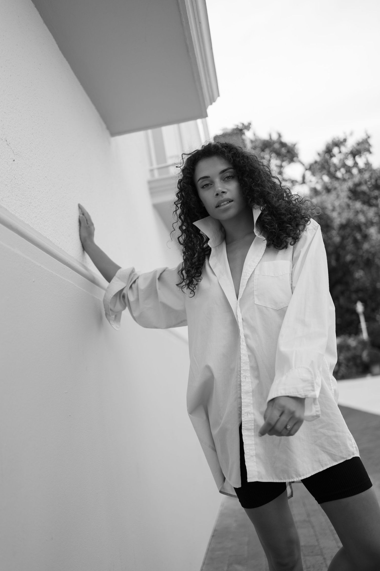 Black and white photo of a curly-haired woman in an oversized white shirt leaning against a wall.