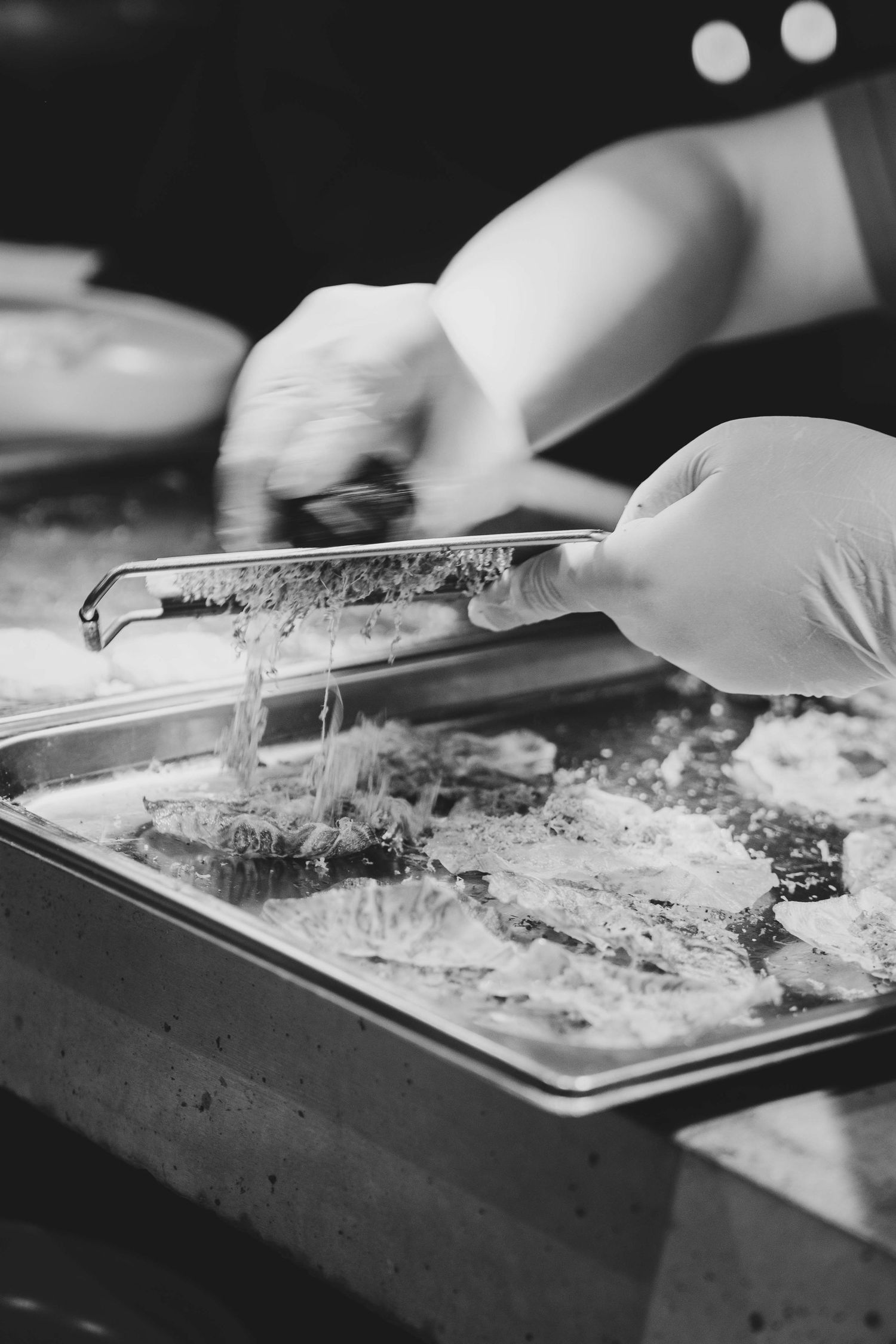 Chef in gloves using tongs to plate cheesy food on a metal tray in a professional kitchen.