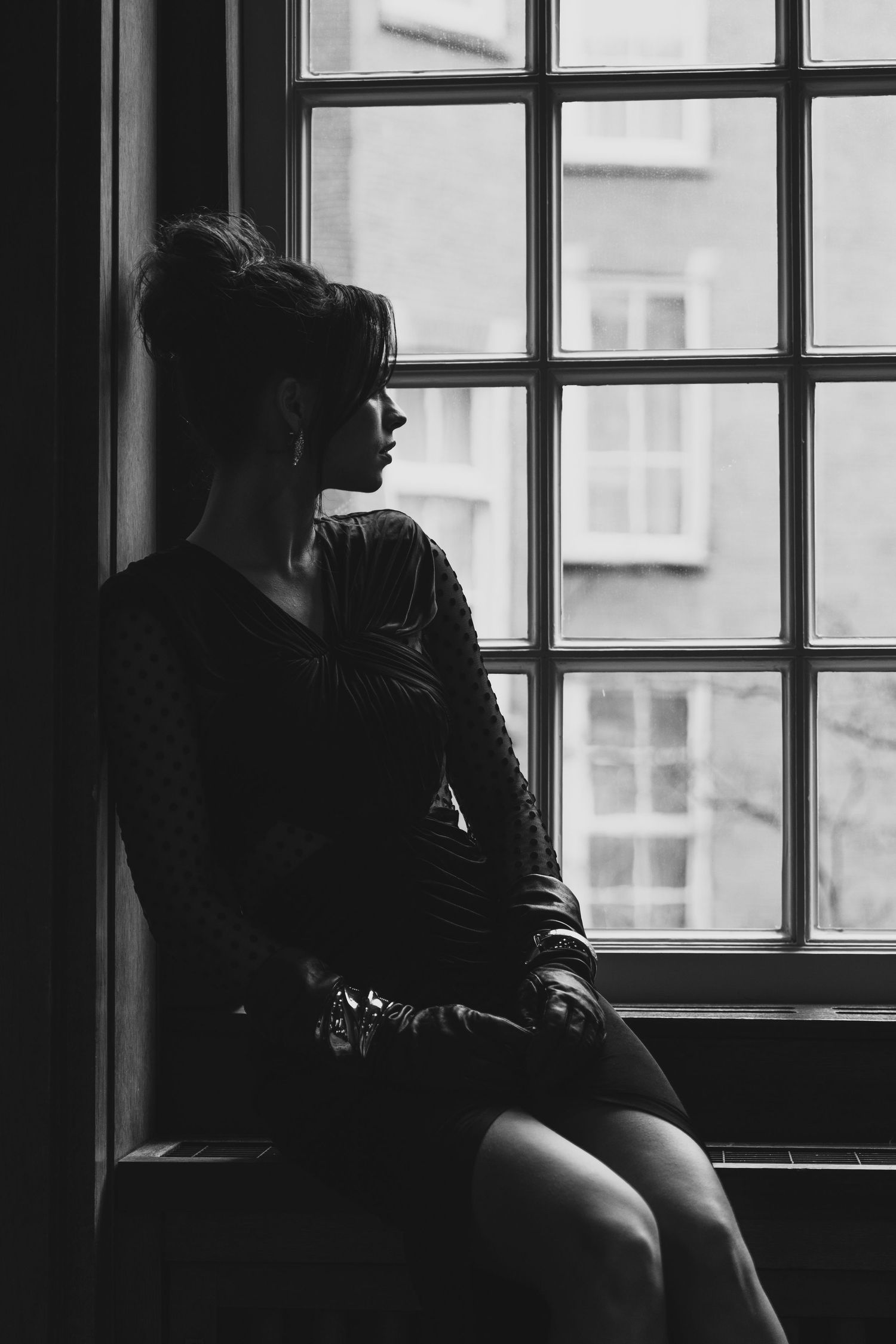 Black and white photo of a woman in a dark dress sitting on a windowsill, gazing outside a large window.