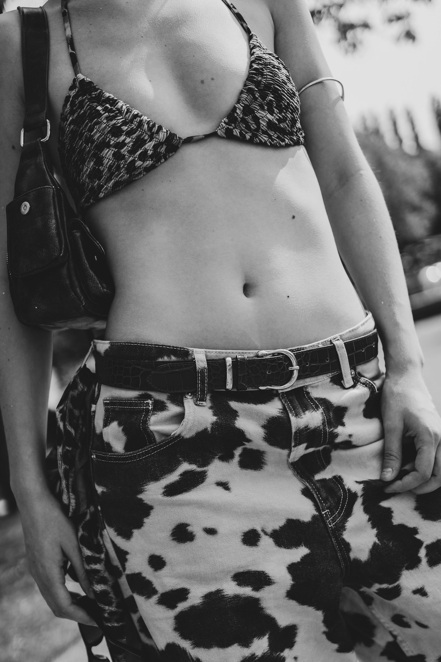 Black and white photo of a woman in a patterned bikini top and cow-print jeans with a belt and crossbody bag.