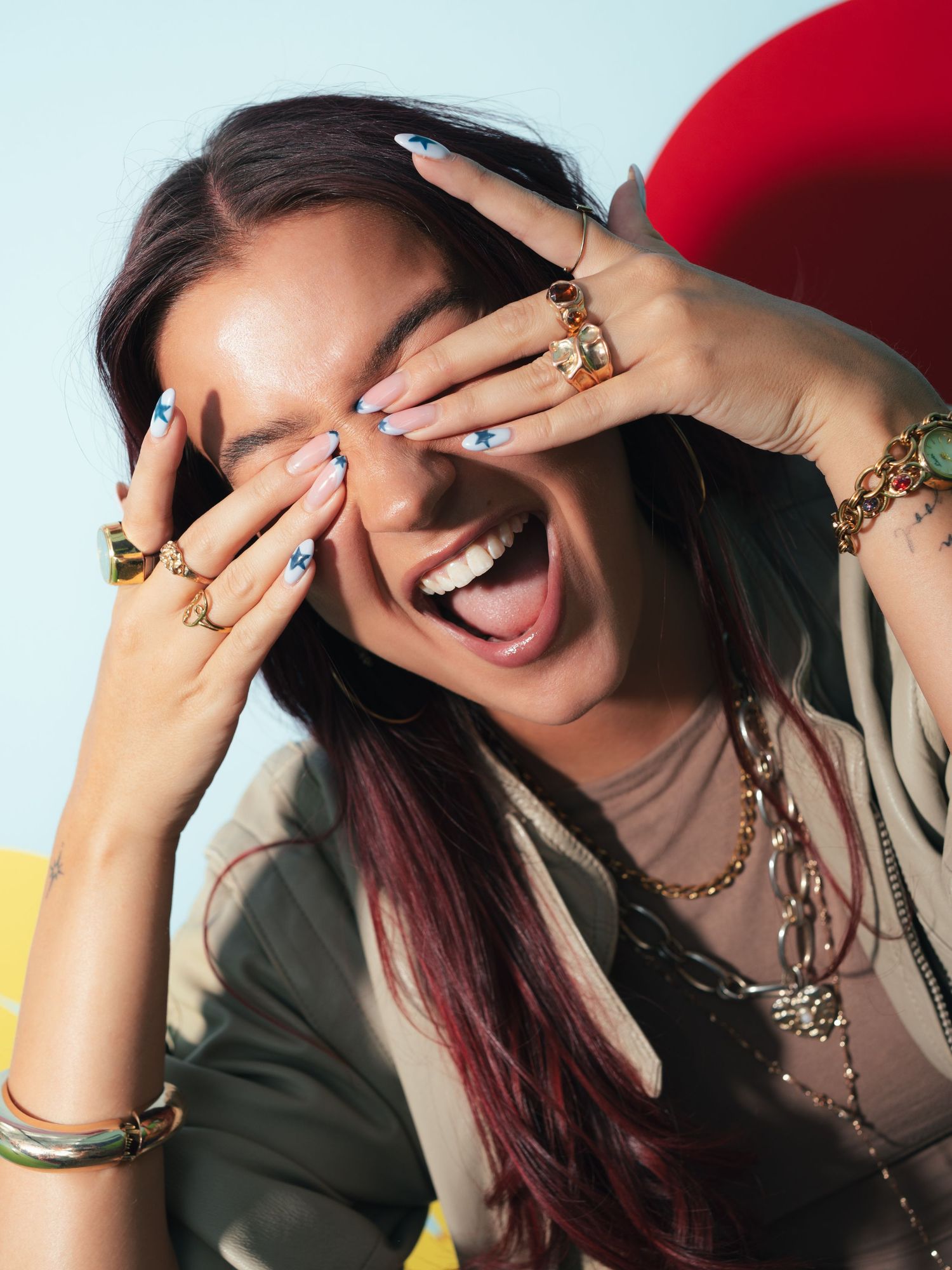 Laughing woman covering eyes with ringed hands, wearing gold jewelry and colorful nail art against a light blue background.